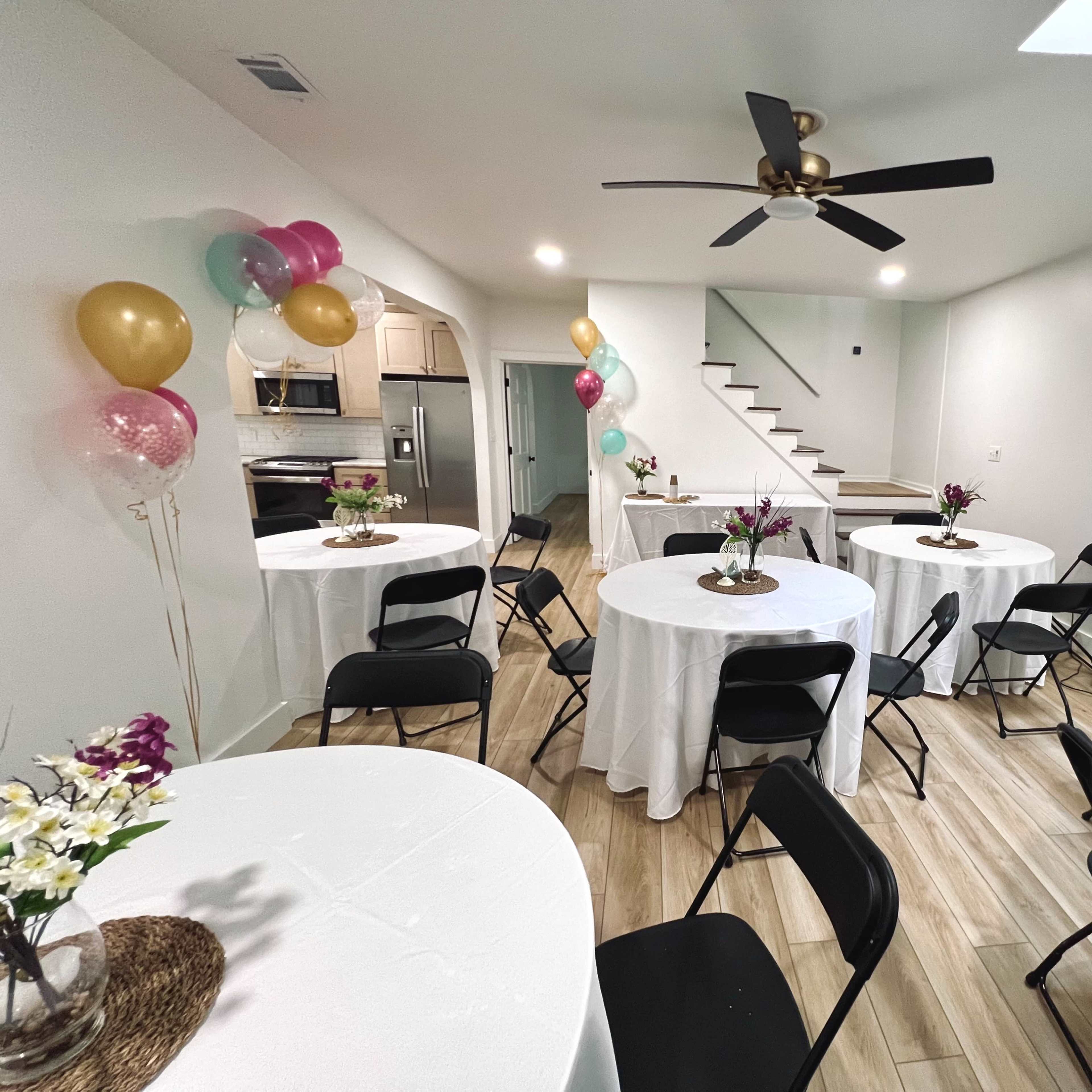 A spacious room features several round tables with white tablecloths, black chairs, and decorative balloons, leading to a staircase at the back.