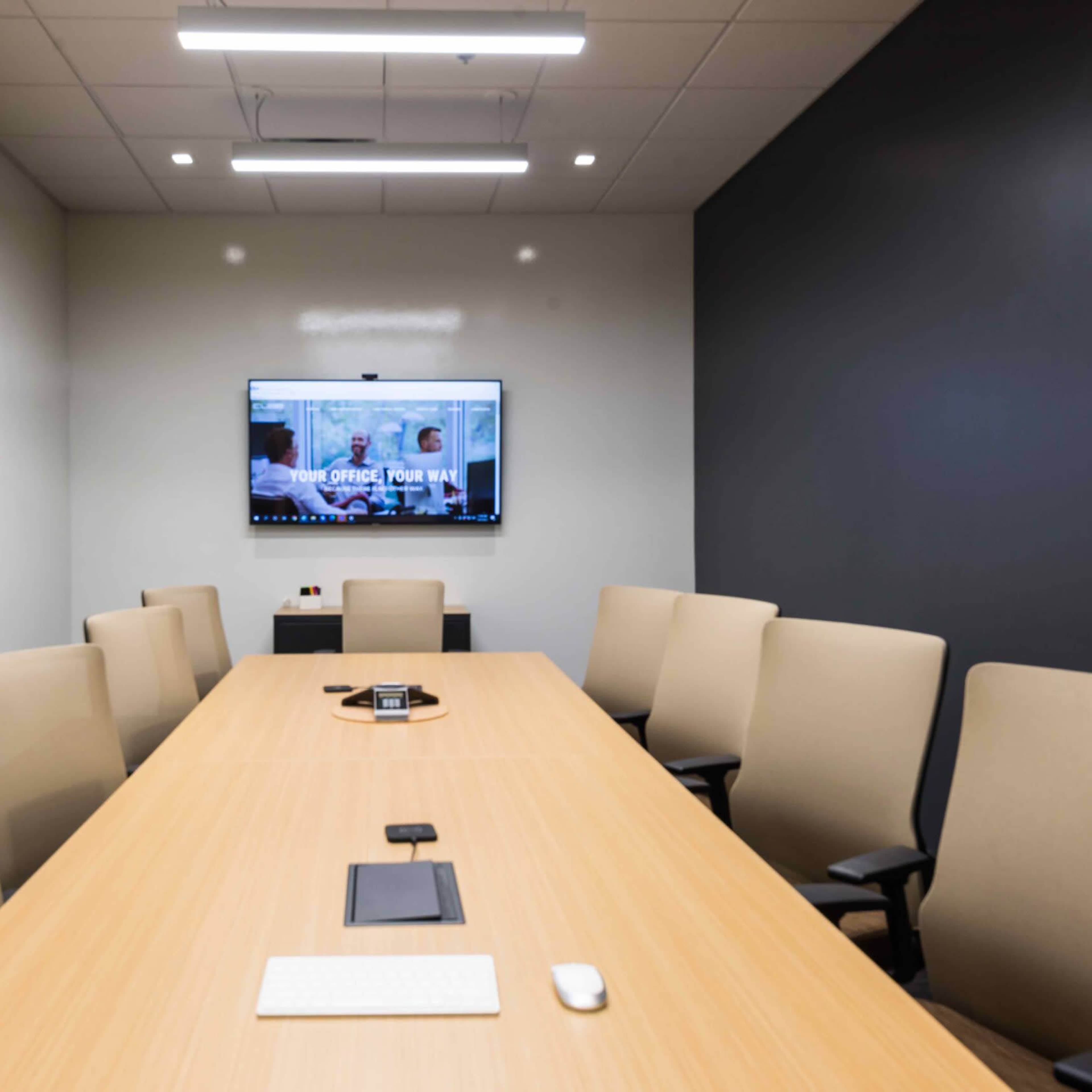 A modern conference room features a long wooden table surrounded by beige chairs, with a large screen mounted on the wall displaying a video call interface.