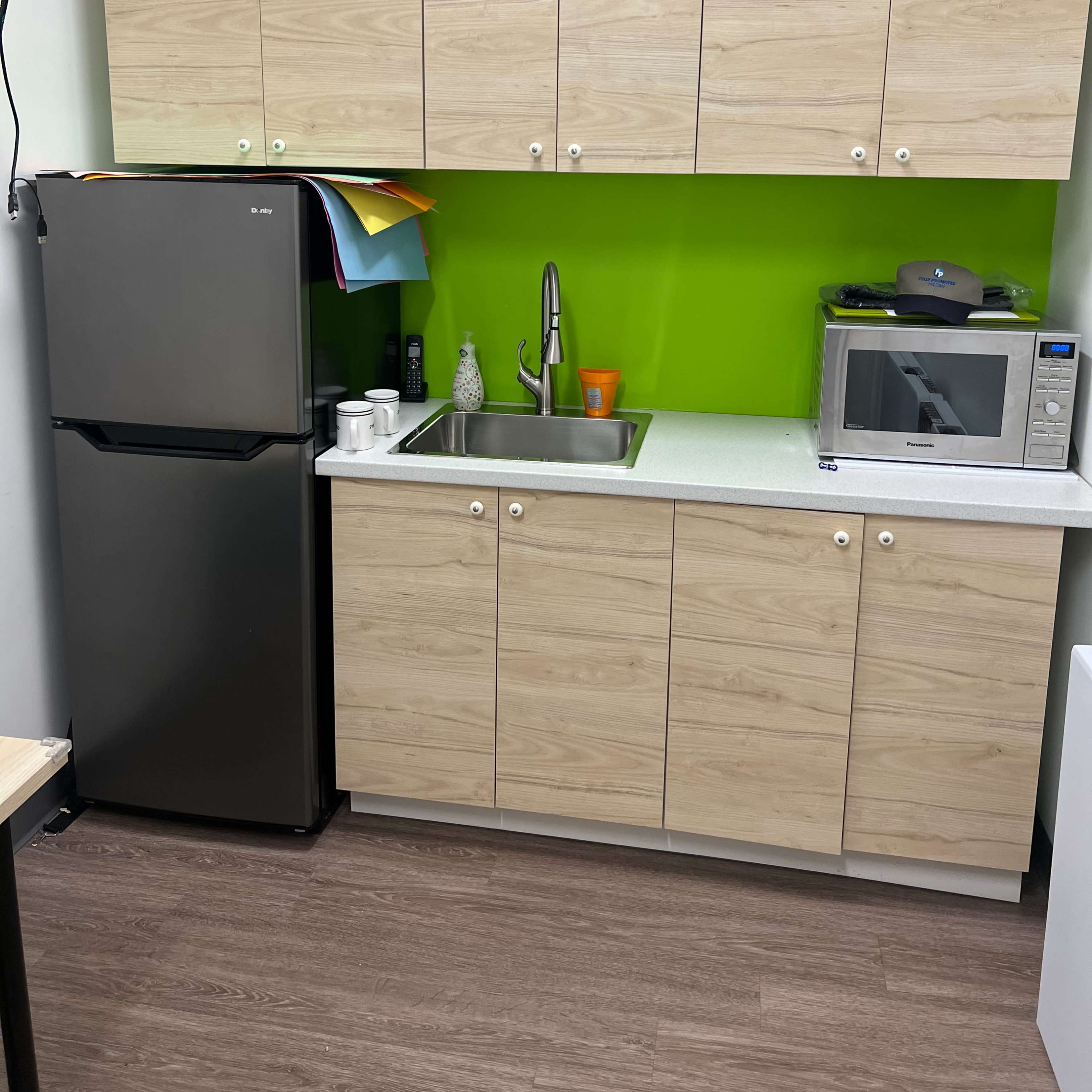 A kitchenette with a black refrigerator, a sink, a microwave, and wooden cabinets above a green wall.