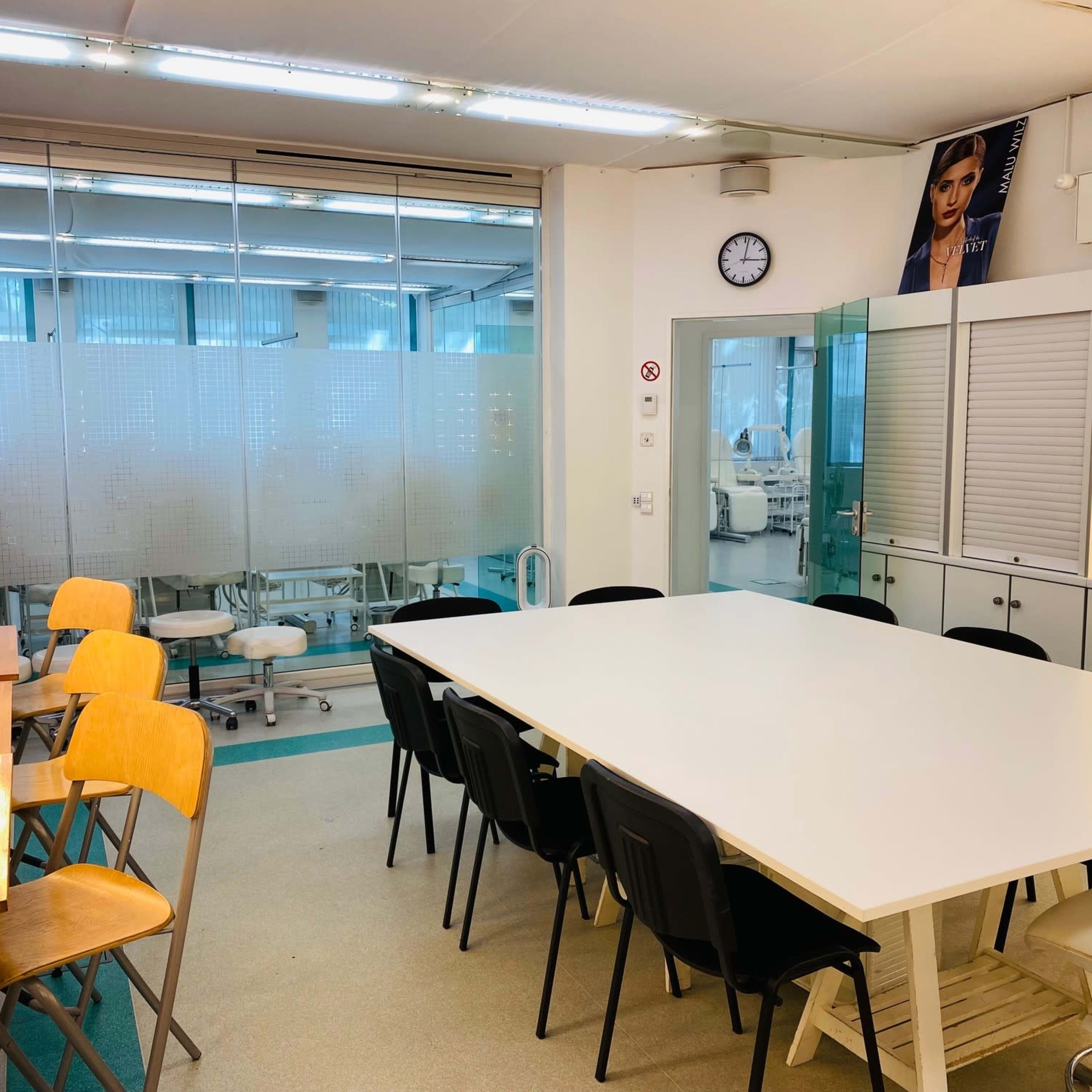 A brightly lit room with a large table and several chairs, alongside a vanity area with mirrors and lights, and a background of medical equipment visible through glass partitions.