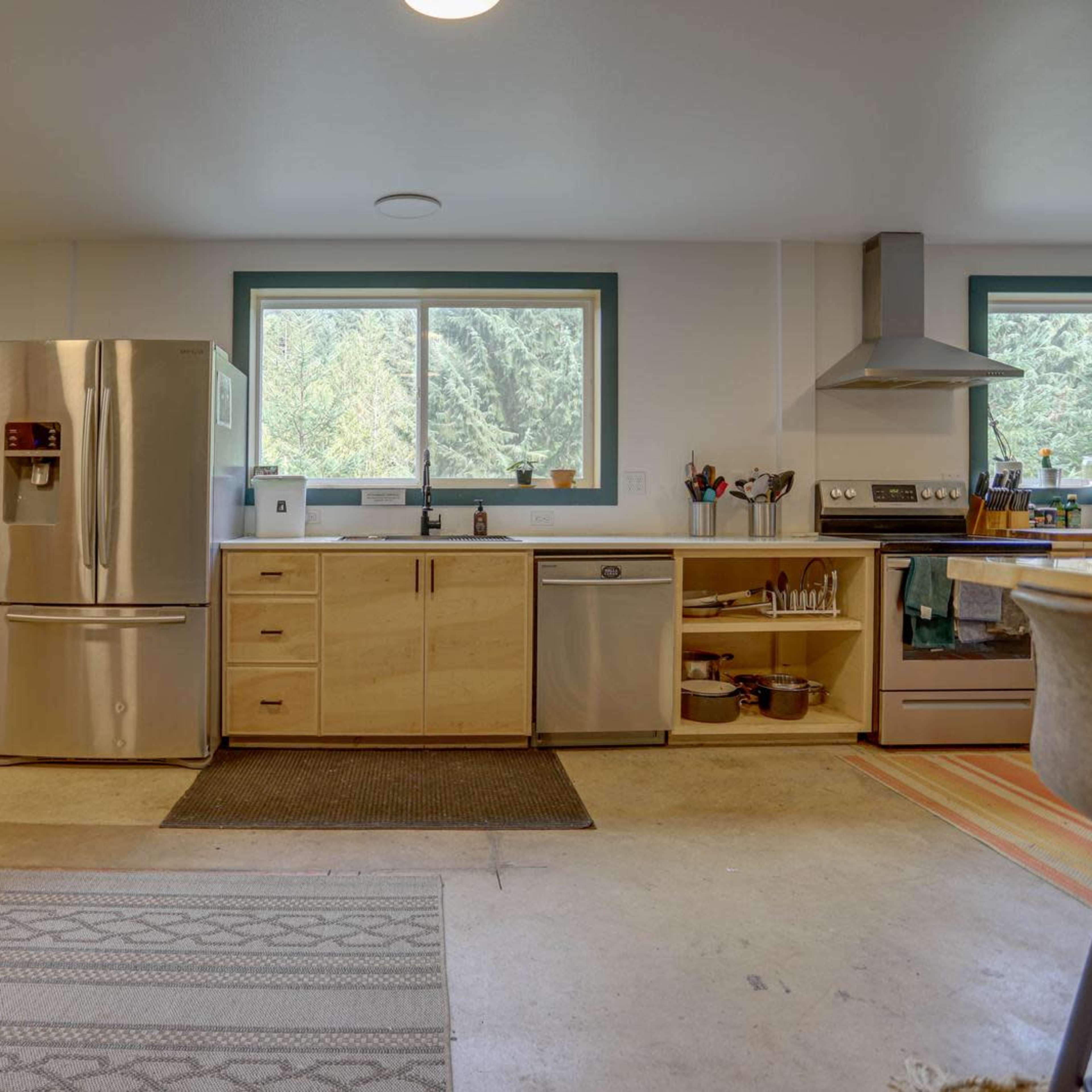 A modern kitchen features a stainless steel refrigerator, an island with seating, and large windows overlooking a wooded area.