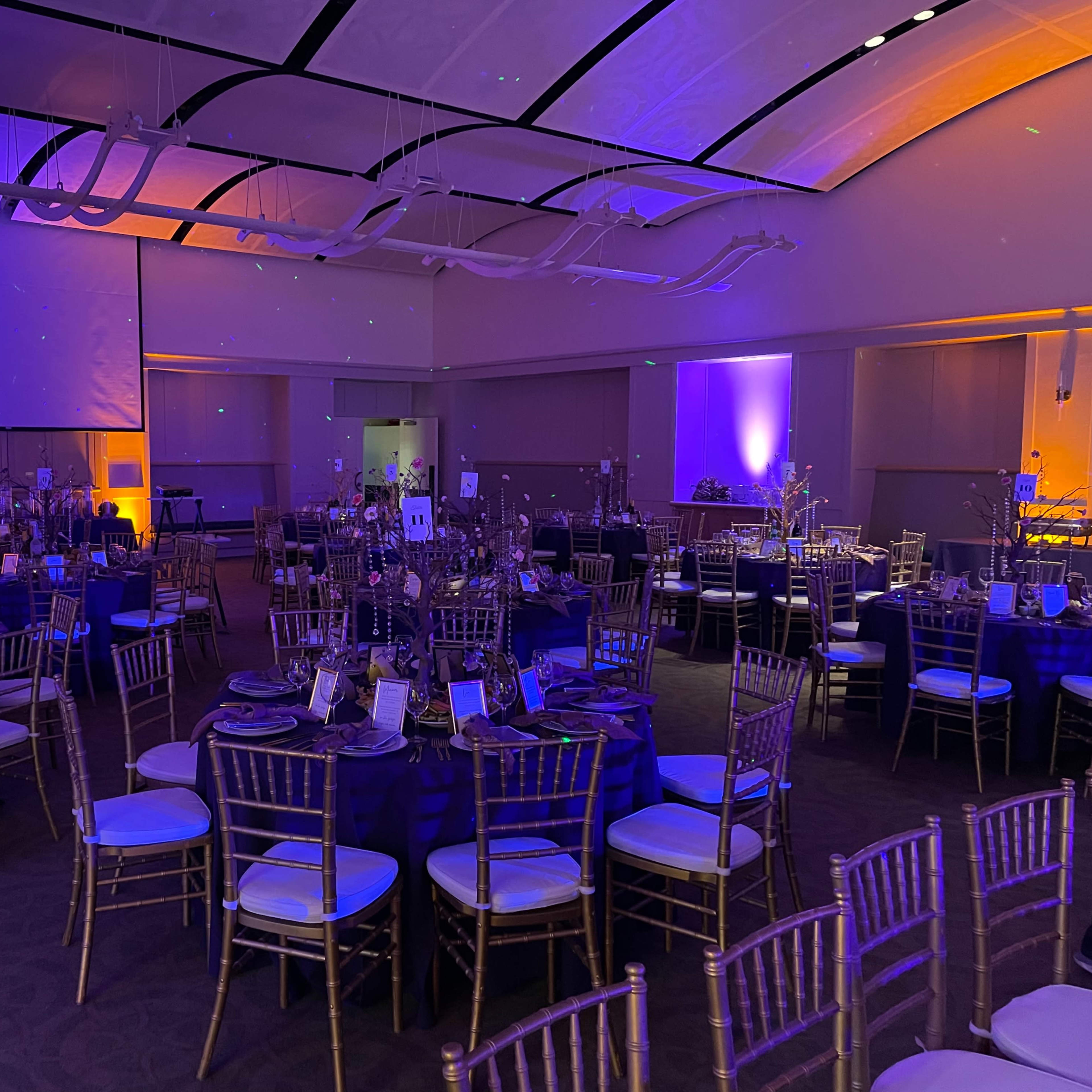 A banquet hall set up for an event, featuring round tables adorned with blue tablecloths and gold chiavari chairs, illuminated by purple and orange lighting.