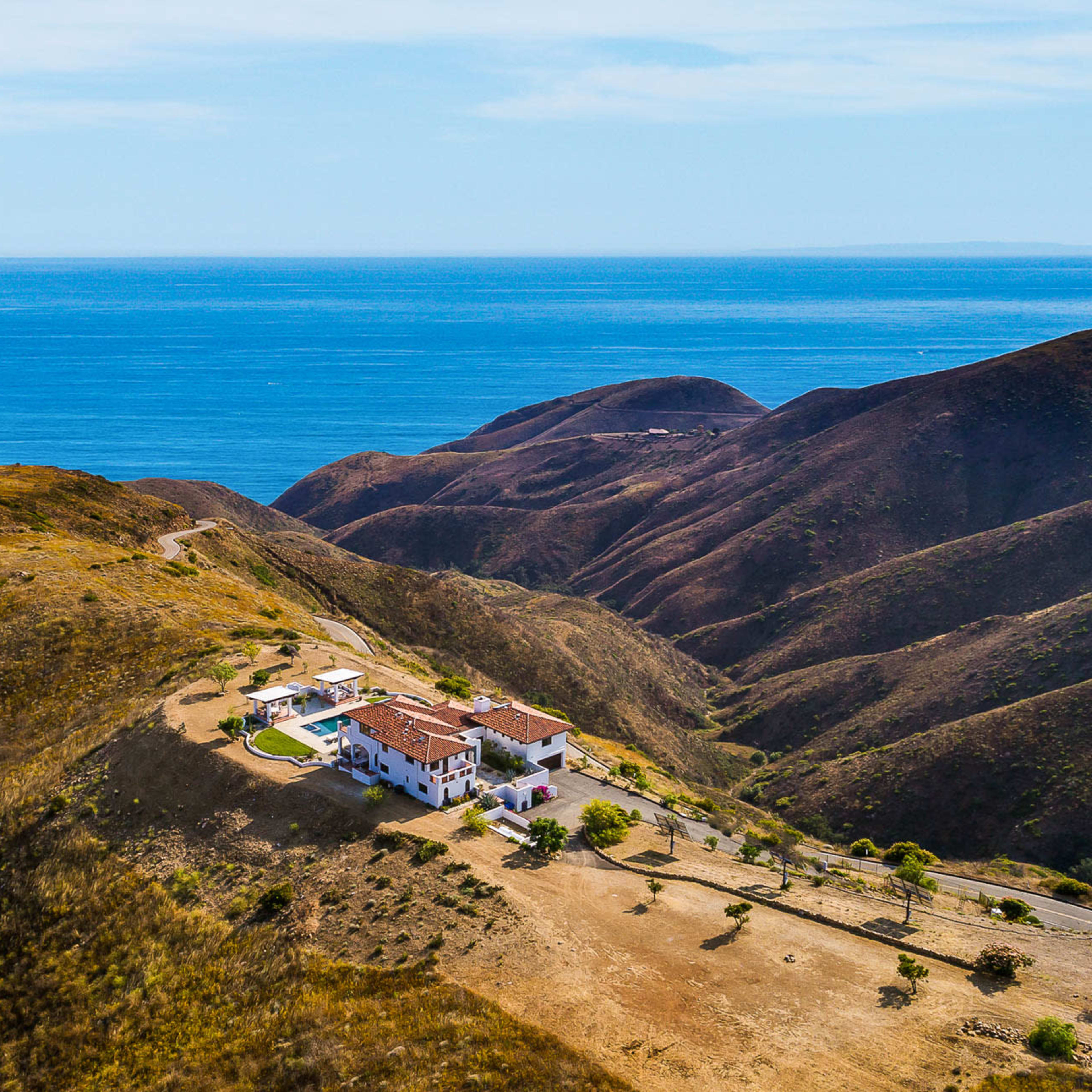A large house sits on a hillside overlooking the ocean, surrounded by rolling brown hills.