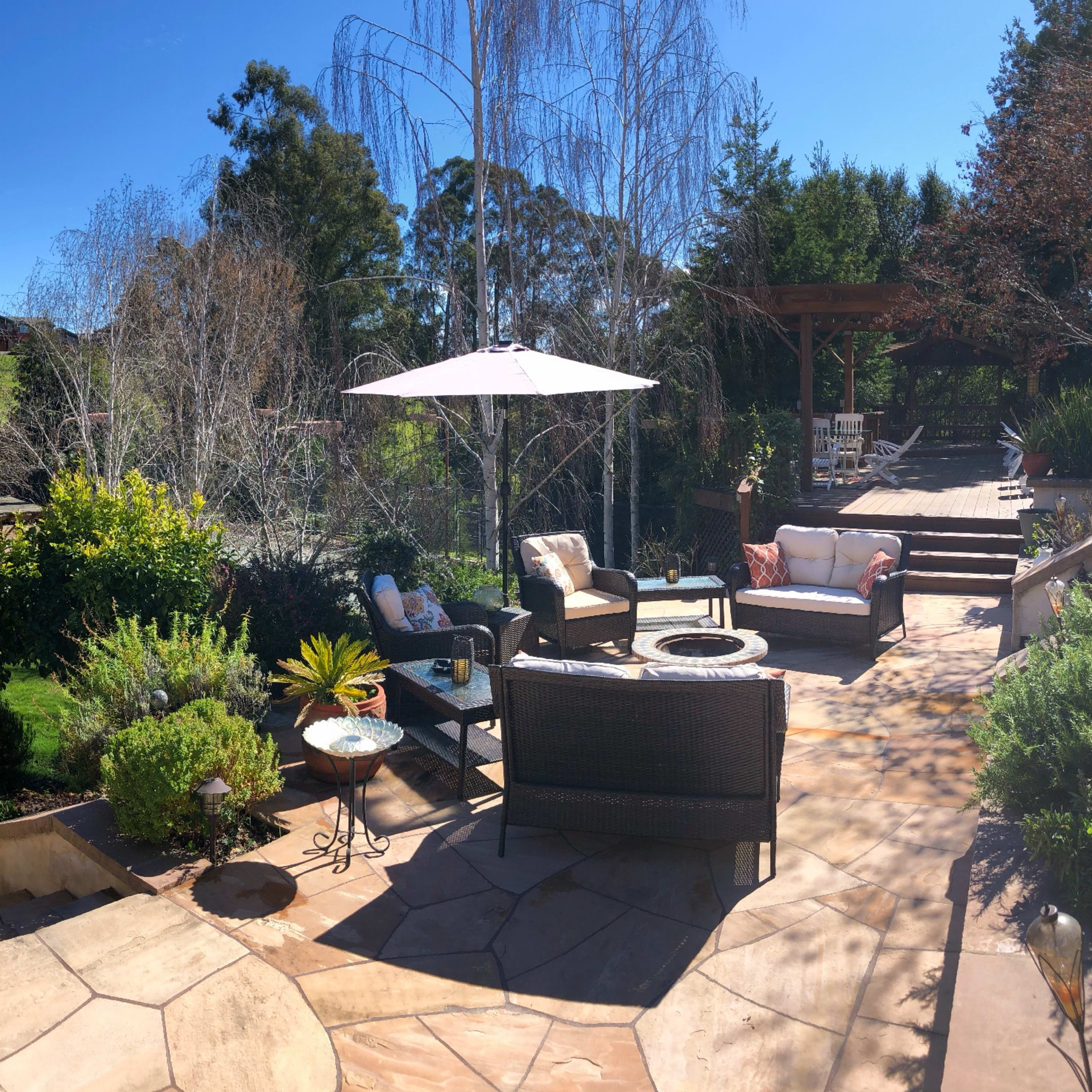 A landscaped outdoor patio area with seating arrangements, surrounding greenery, and a patio umbrella under a clear blue sky.