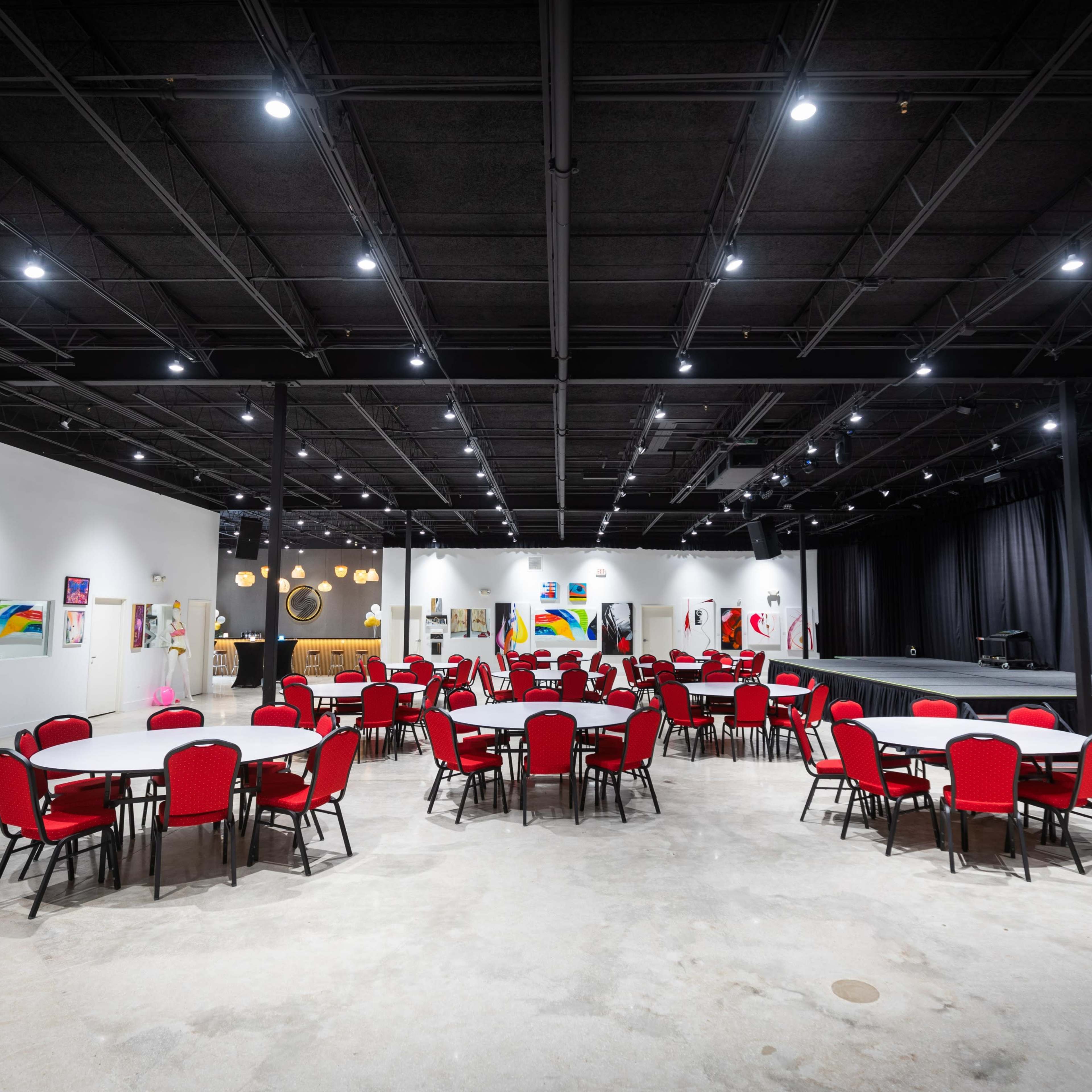 A large, empty event space with circular tables and red chairs arranged around them, and a stage at the far end.