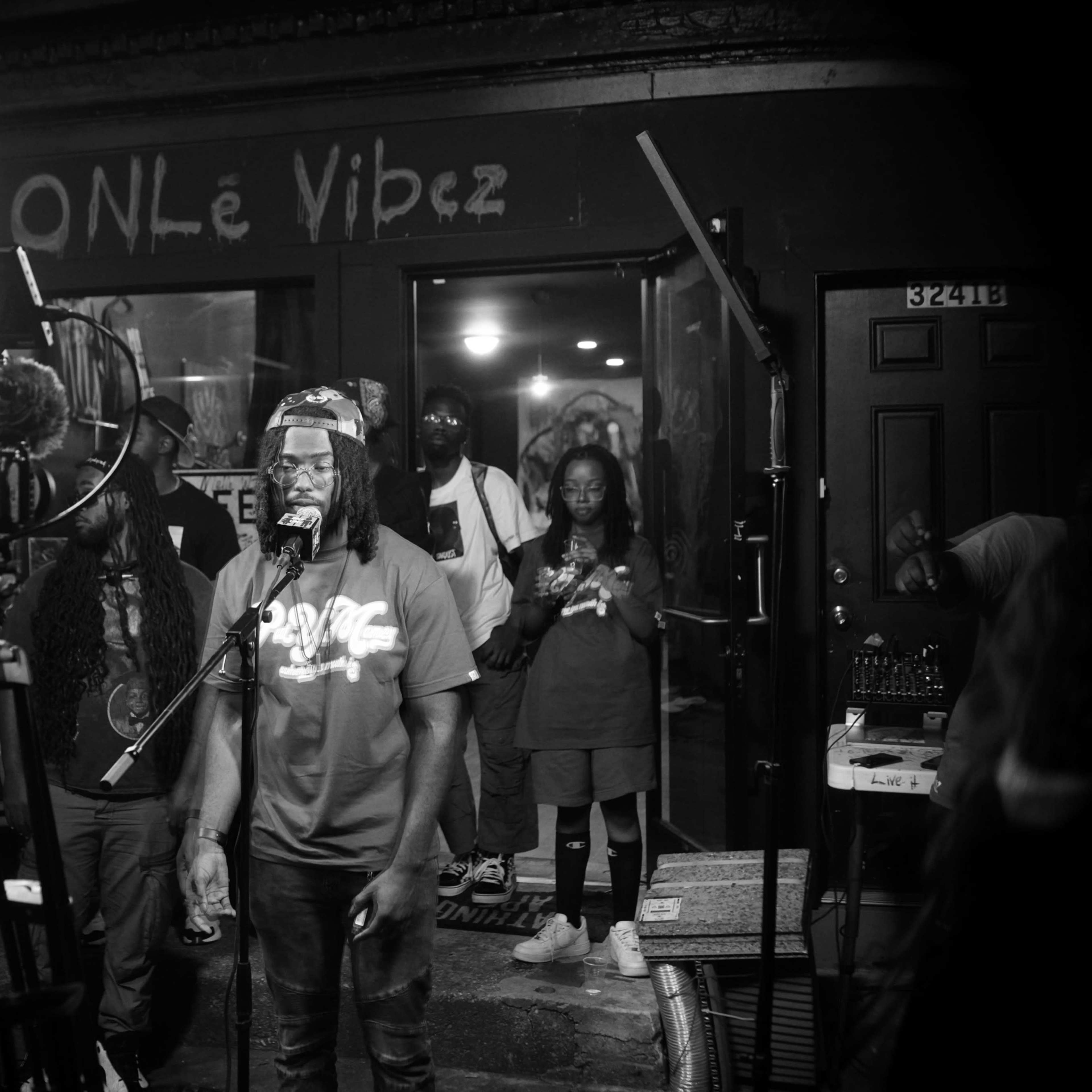 A group of performers and audience members gathers outside a venue called "ONLe Vibez," where one musician is speaking into a microphone.