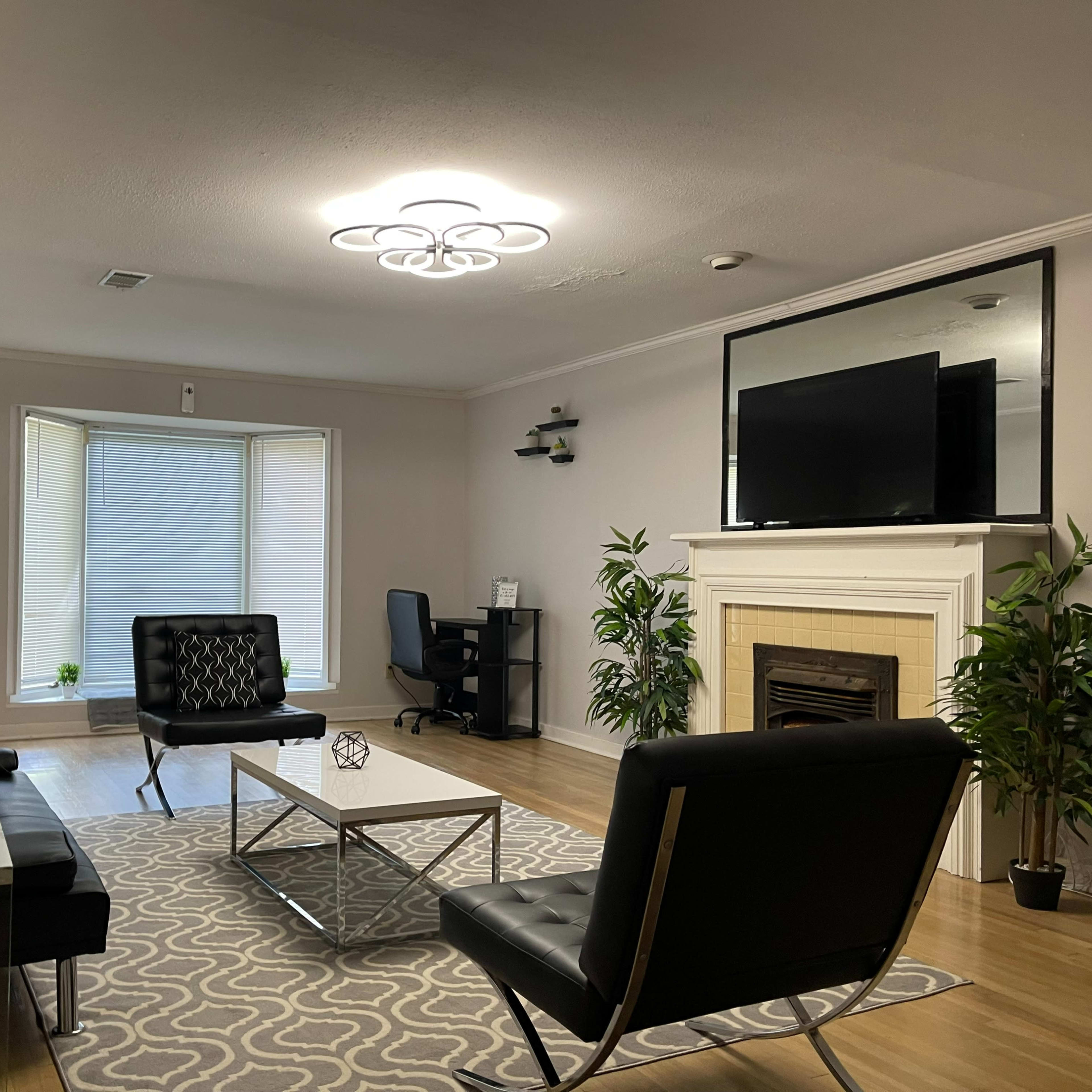 A modern living room featuring a black sofa set, a coffee table on a patterned rug, a fireplace, and a television above it, with a desk in the corner.