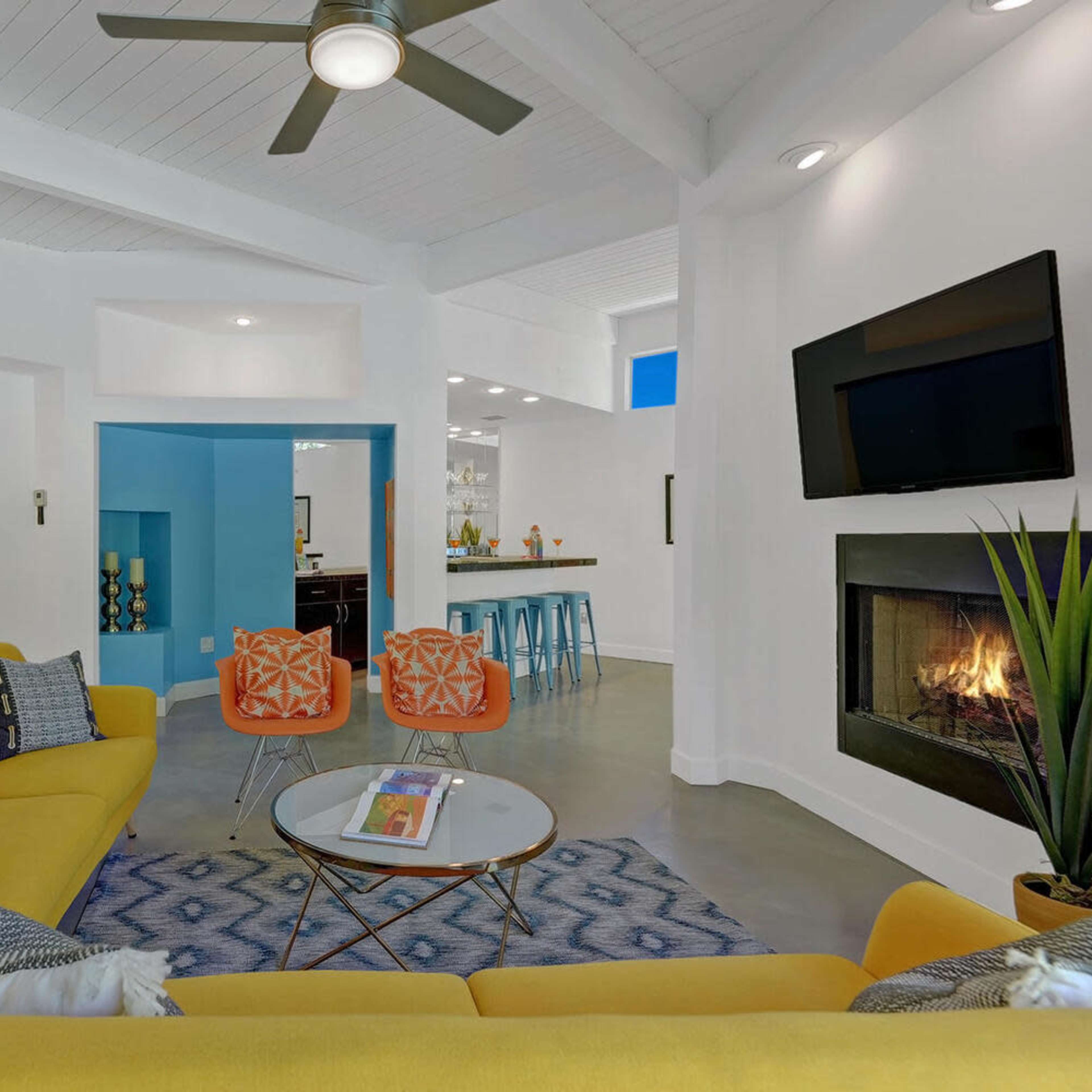 A modern living room featuring a yellow sectional sofa, a glass coffee table, a fireplace, and an open layout leading to a kitchen area with bar stools.
