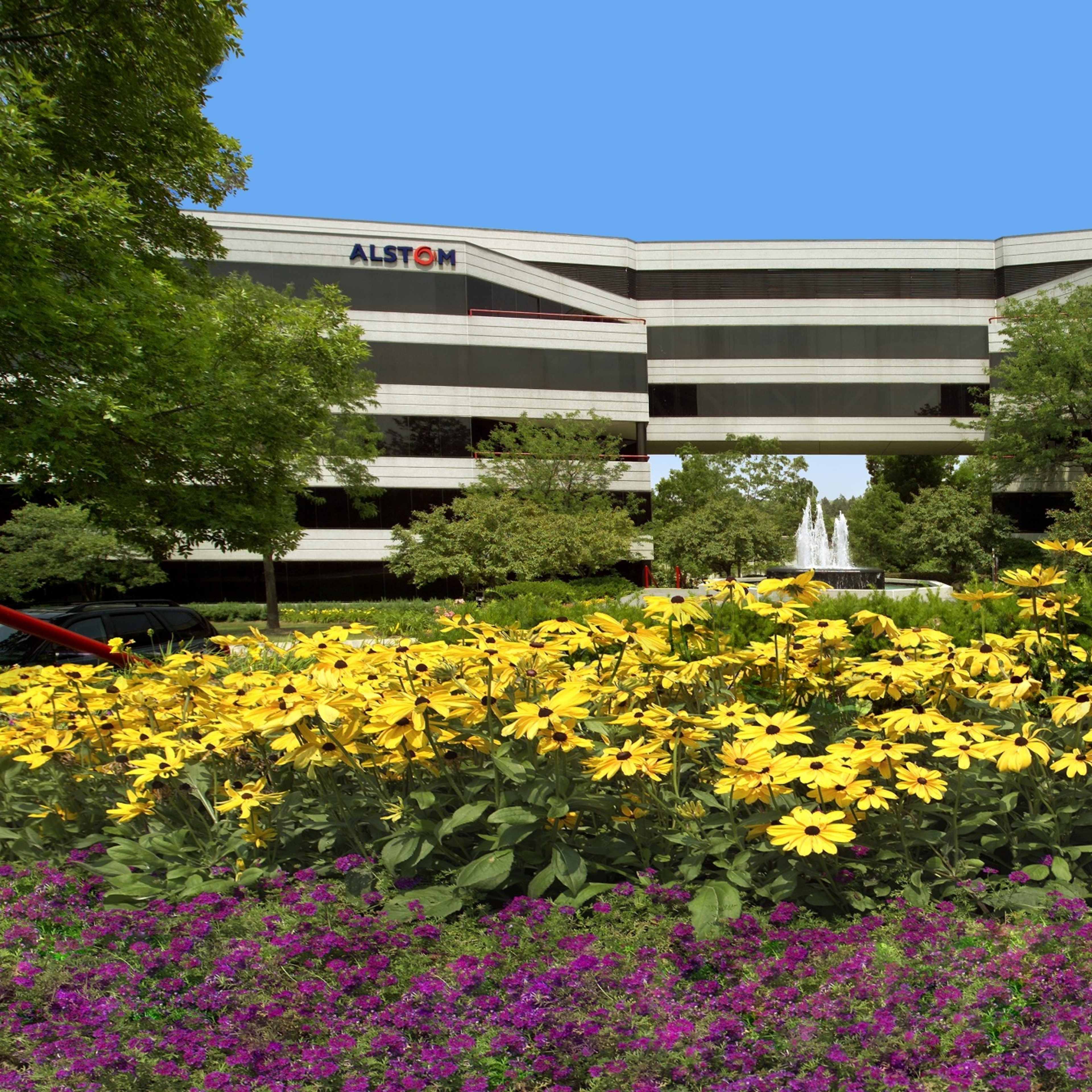 A modern office building with the Alstom logo is framed by vibrant flower beds and trees in a landscaped area.