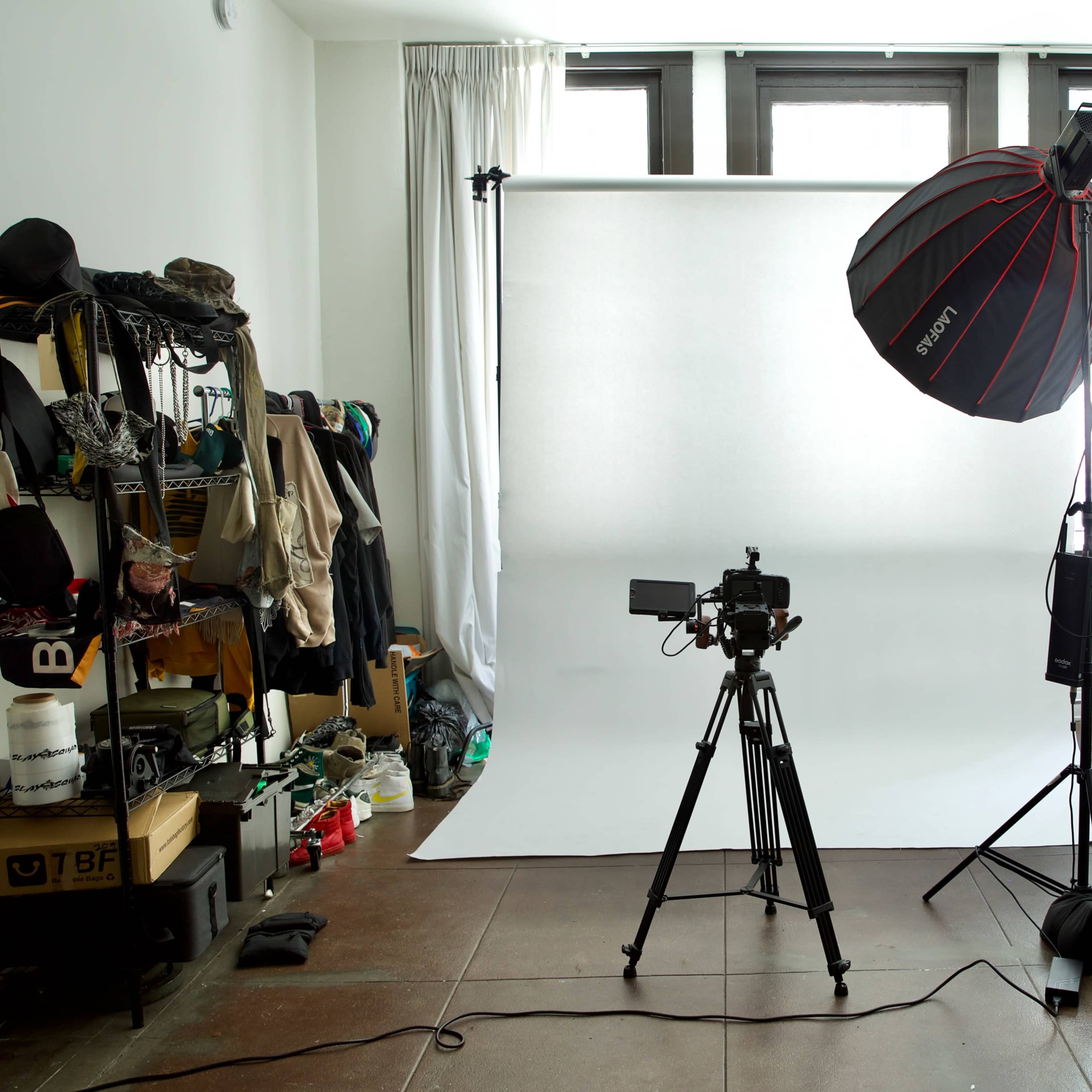 A photography setup featuring a camera on a tripod, a large softbox light, and a white backdrop in a room with various personal items and a bed visible in the background.