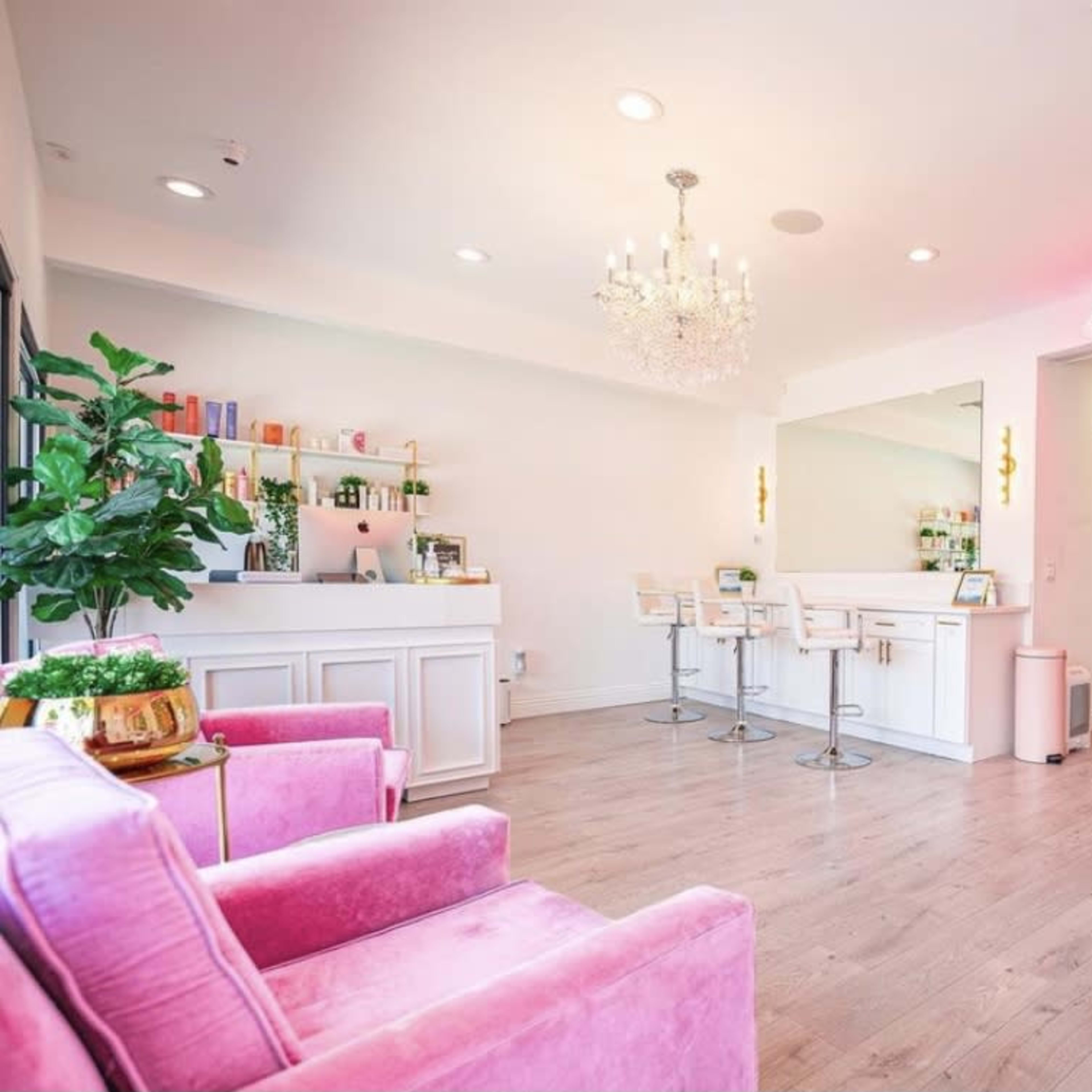A brightly lit beauty salon features pink furniture, a reception area with a bar, and a decorative green wall.