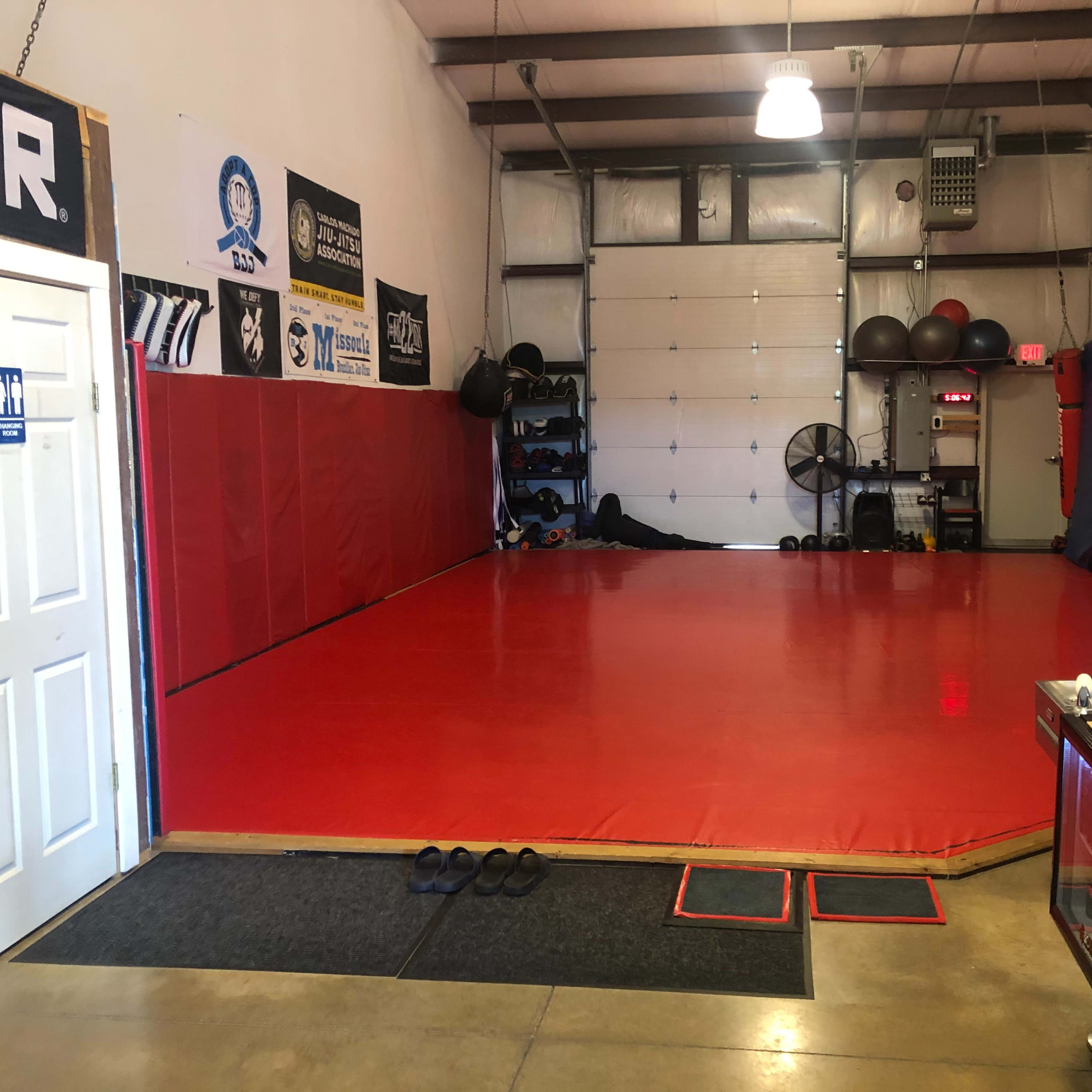 A martial arts training facility with a red mat, gym equipment along the walls, and a door leading to a restroom.