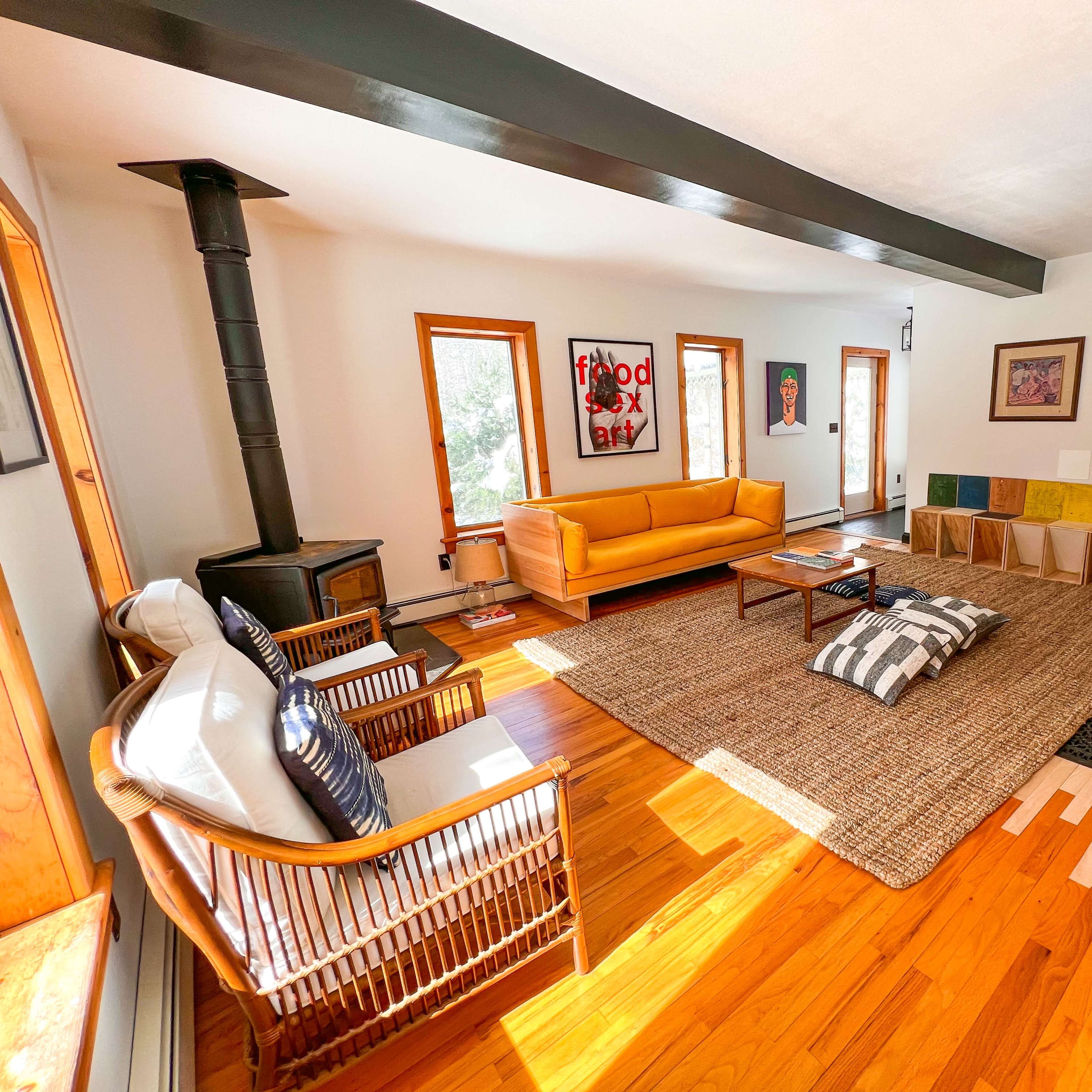A spacious living room with a yellow sofa, a wood-burning stove, and a rug surrounded by several cushions and wooden decor.
