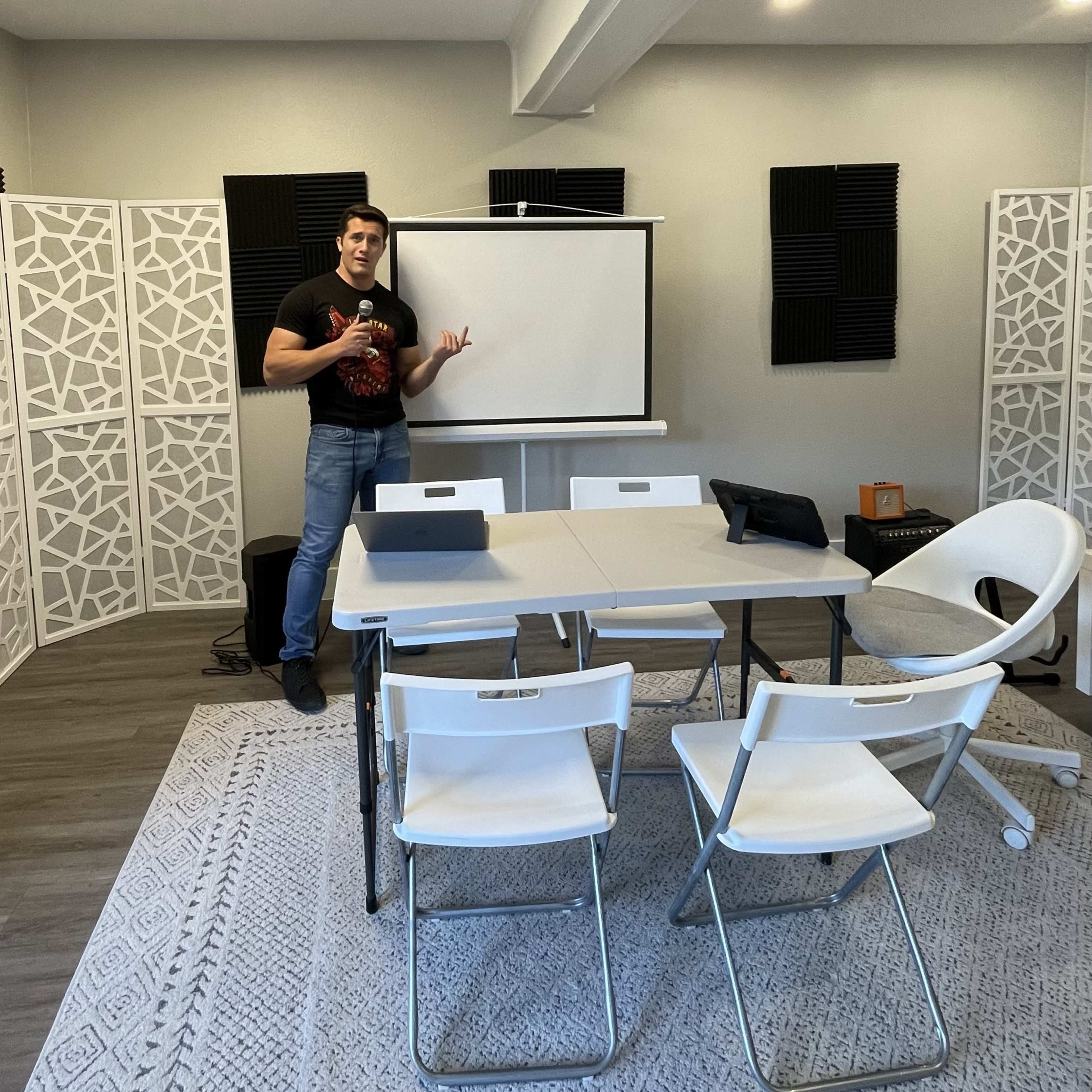 A man stands beside a projector screen in a meeting room furnished with a table and four chairs.
