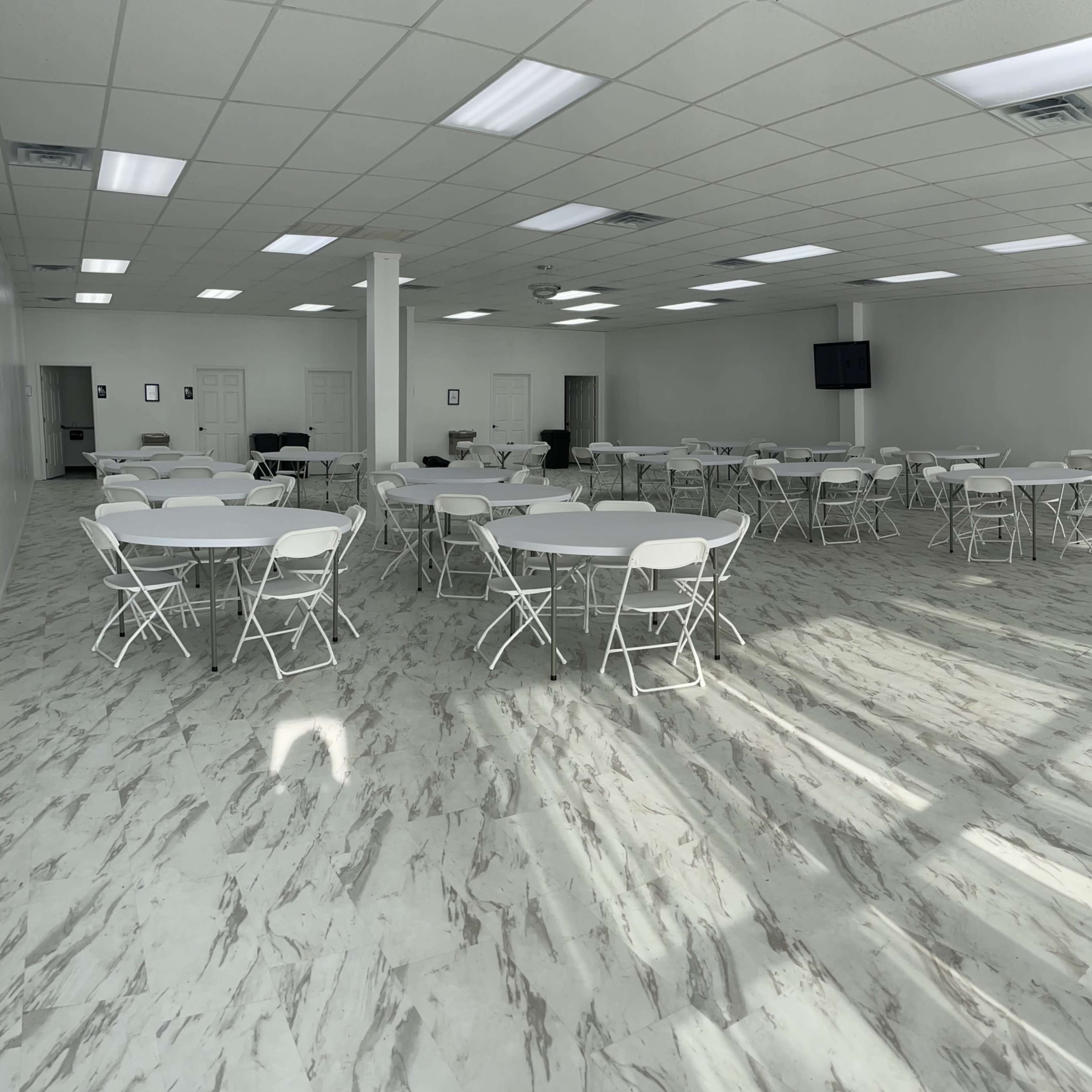 A spacious event room with multiple round tables and chairs arranged neatly, featuring a decorative green wall on one side and sunlight casting shadows on the marble floor.