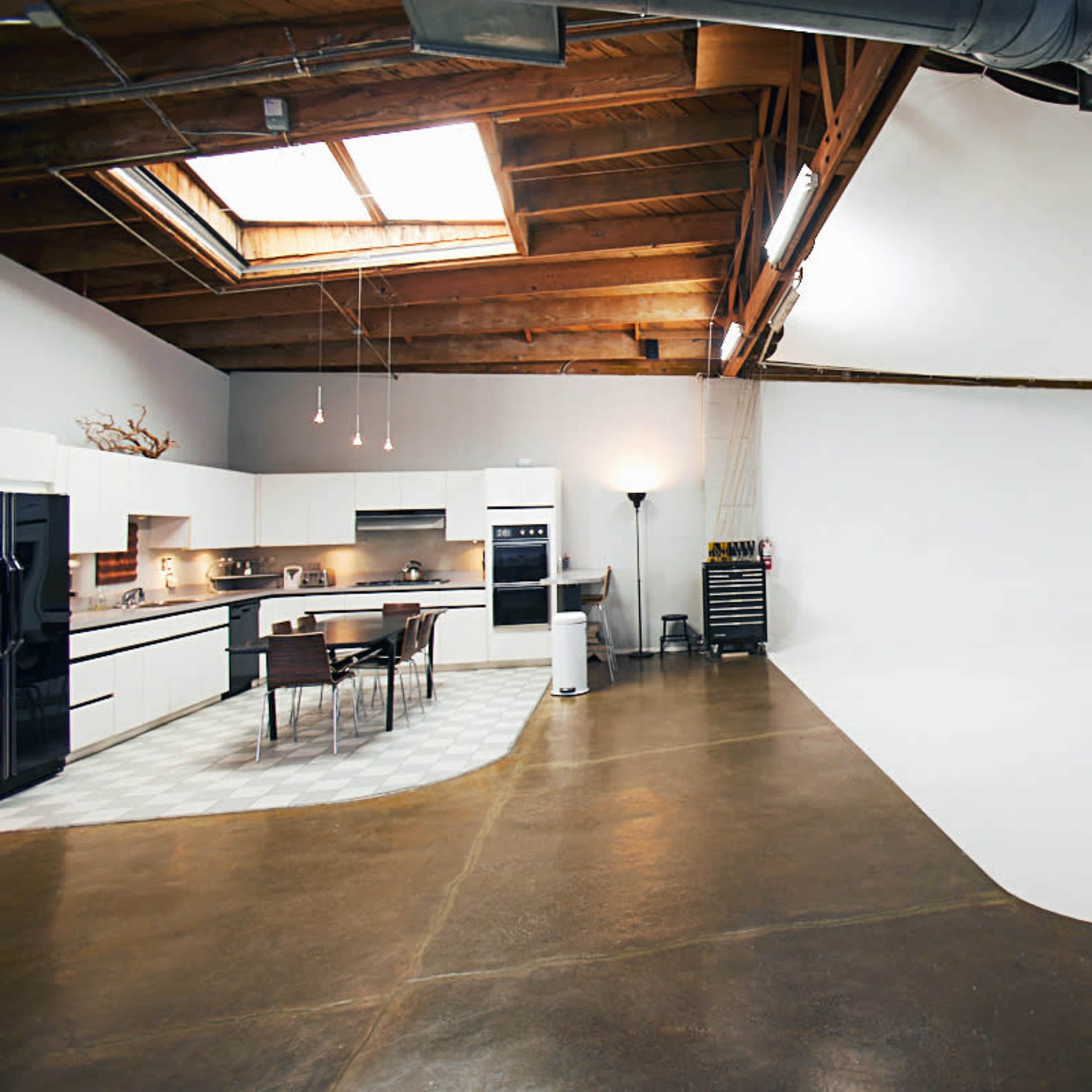 A spacious kitchen with modern appliances, a dining table, and a large white backdrop in an open area with wooden beams and a skylight.