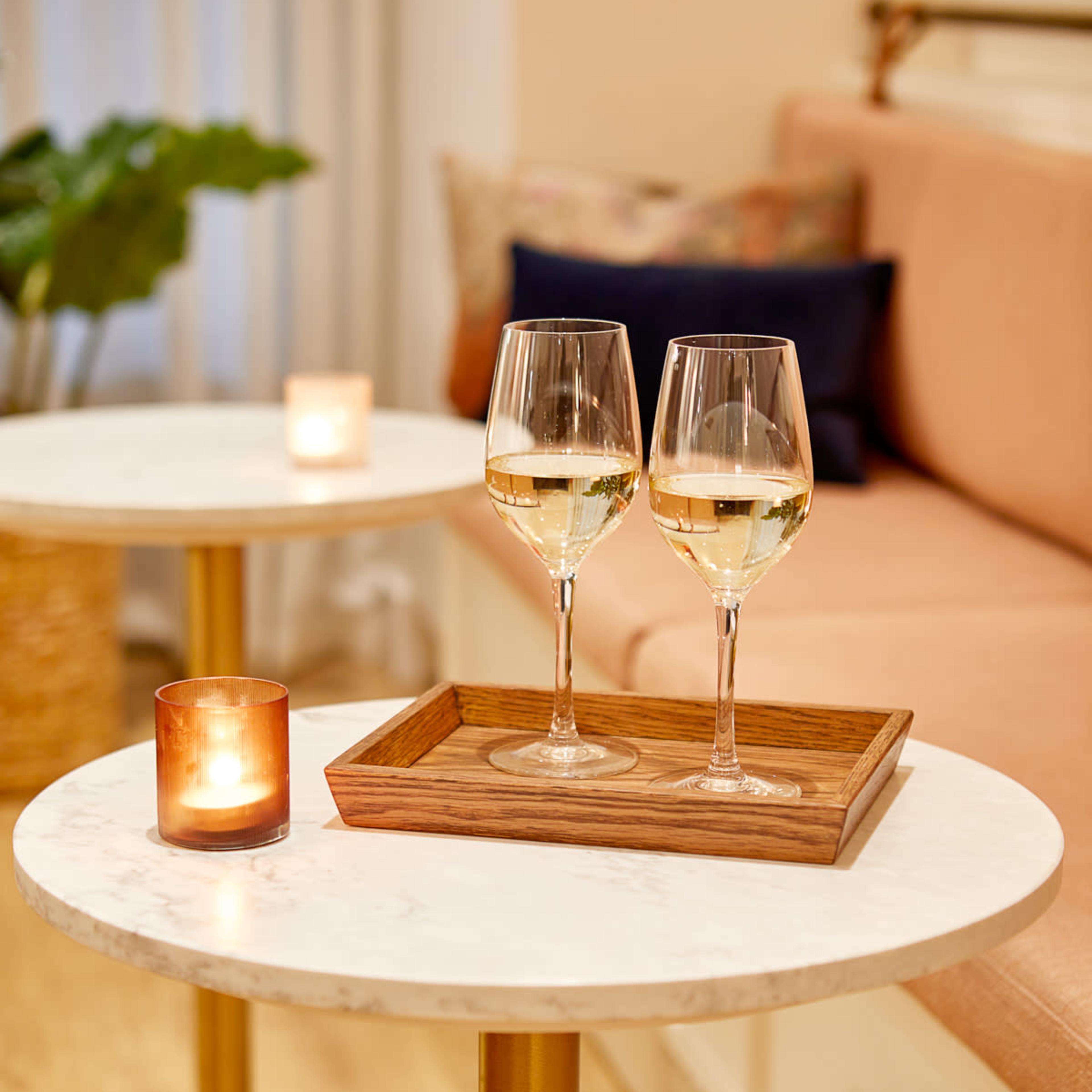 Two glasses of white wine are placed on a wooden tray atop a round marble table beside a lit candle, in a cozy indoor setting.
