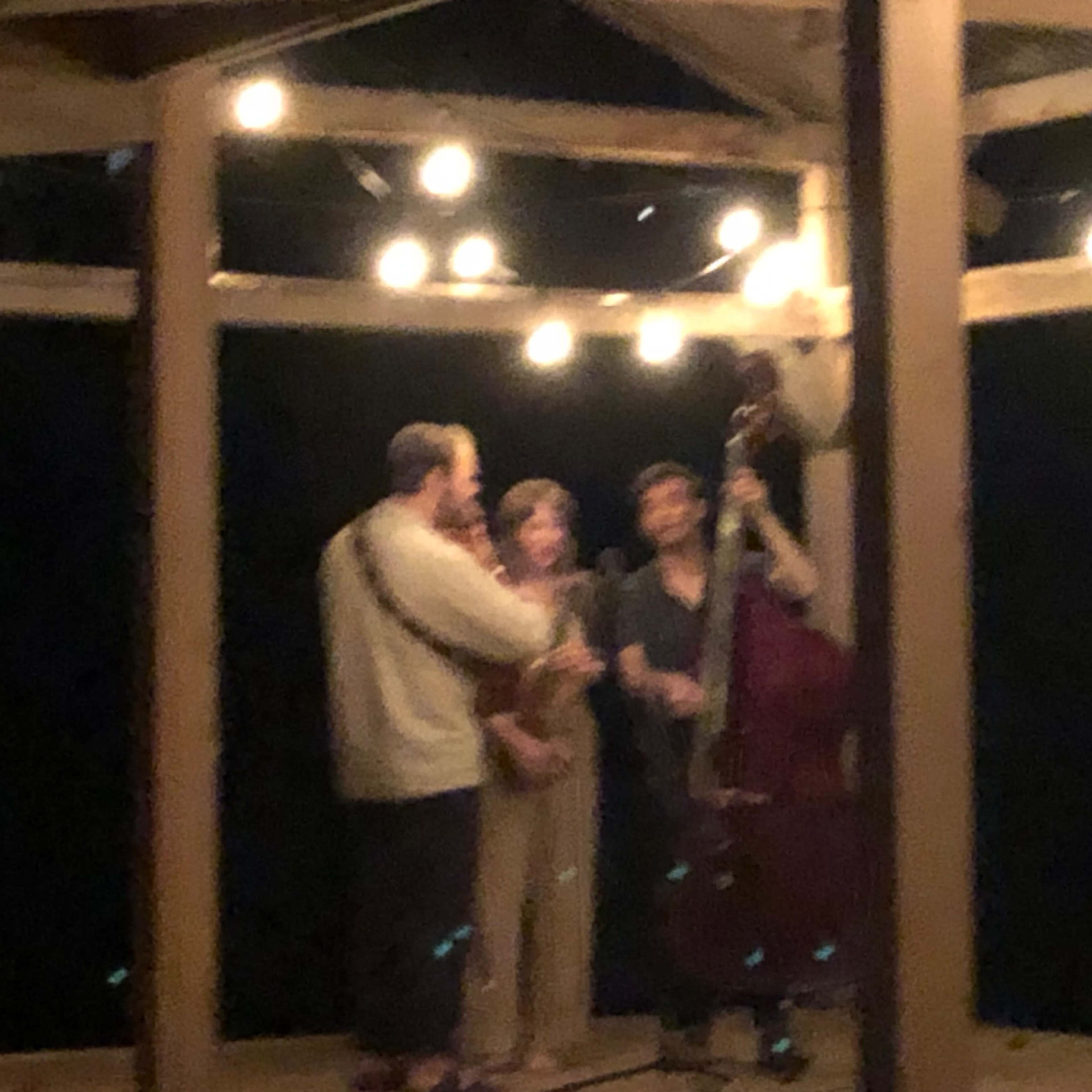 Three musicians perform on a wooden stage beneath string lights at night, with one playing guitar and another on upright bass.