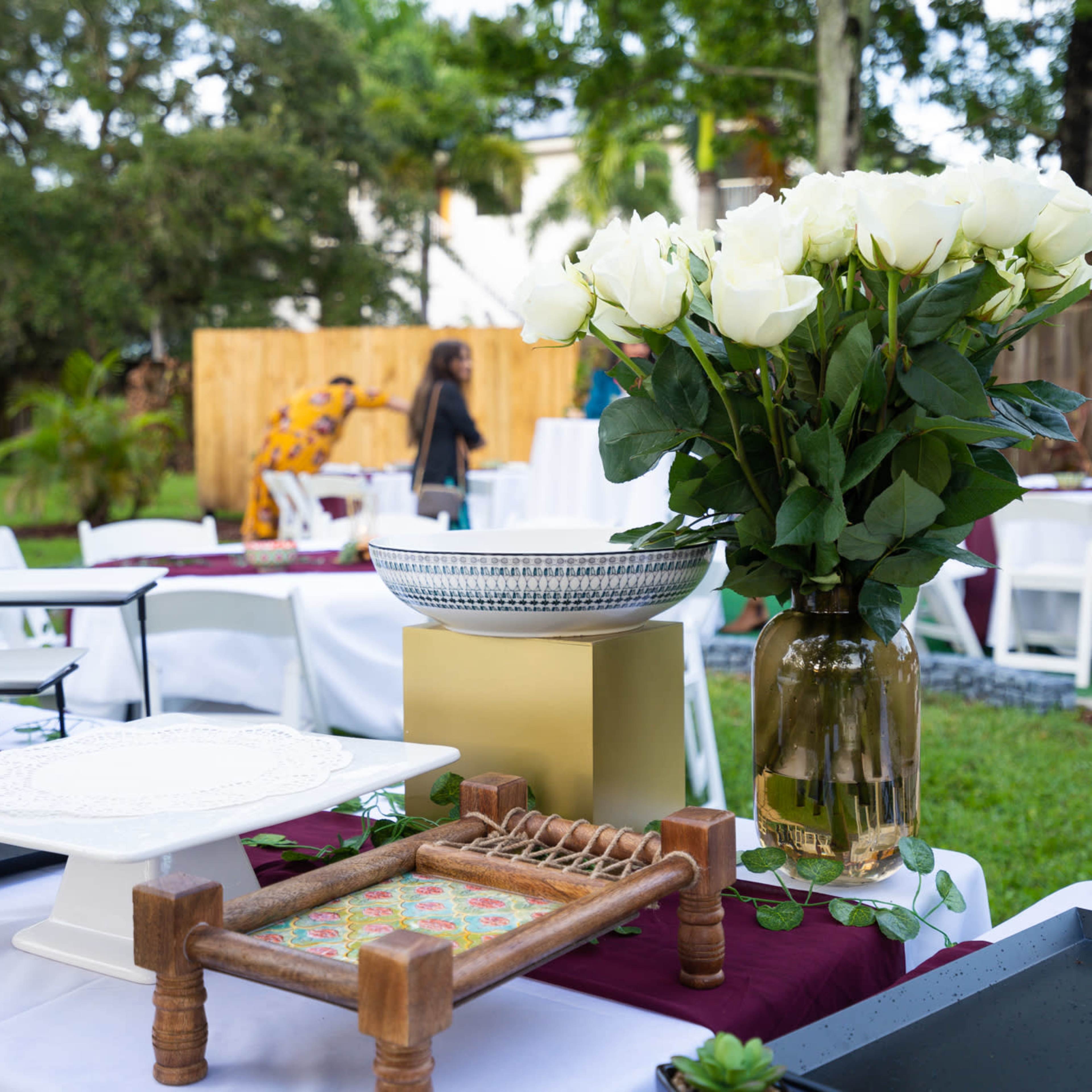 A decorated outdoor setting with white tables, floral arrangements, and various serving trays arranged for an event.