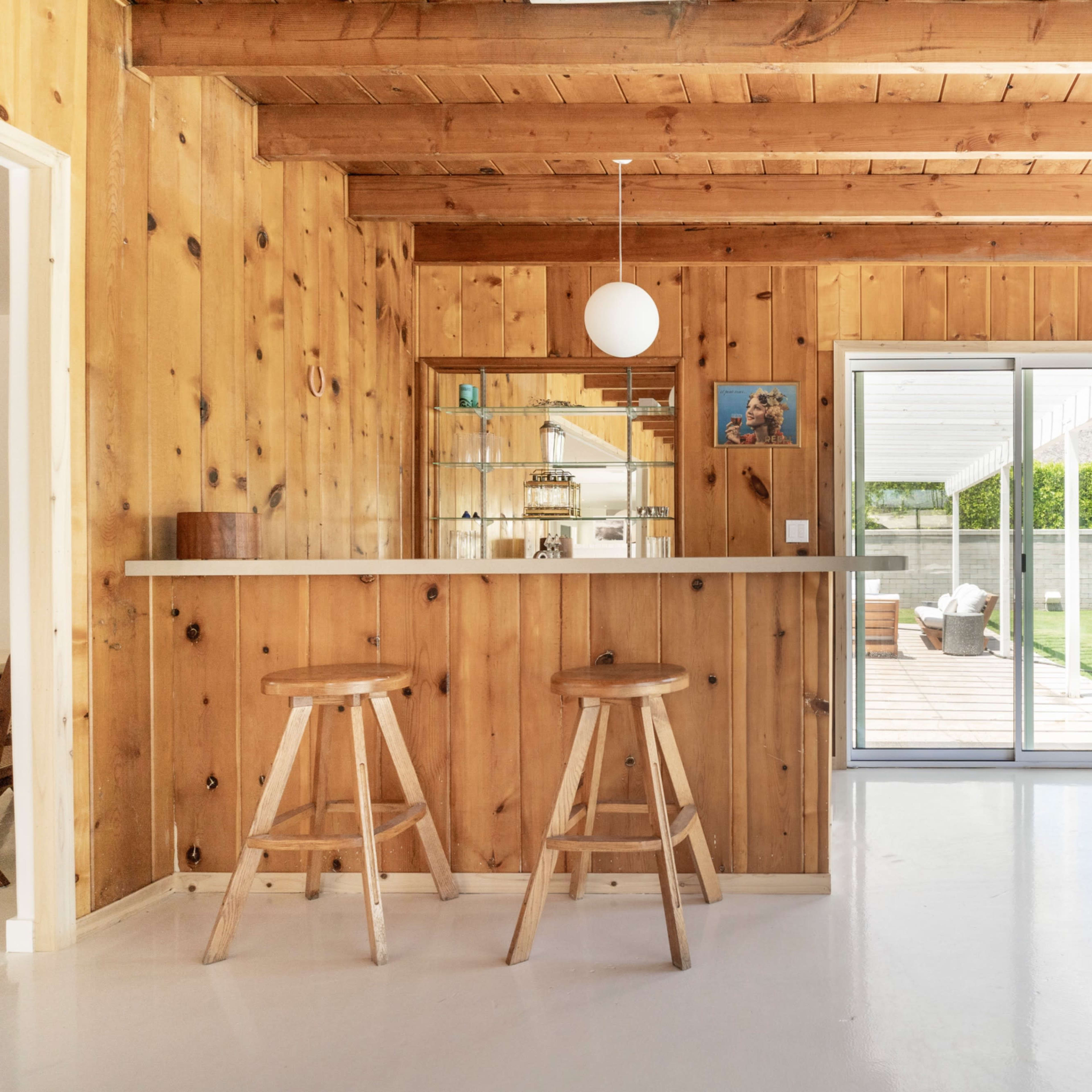 A wooden-paneled interior featuring a bar with two stools, a glass sliding door, and a view of an outdoor patio area.