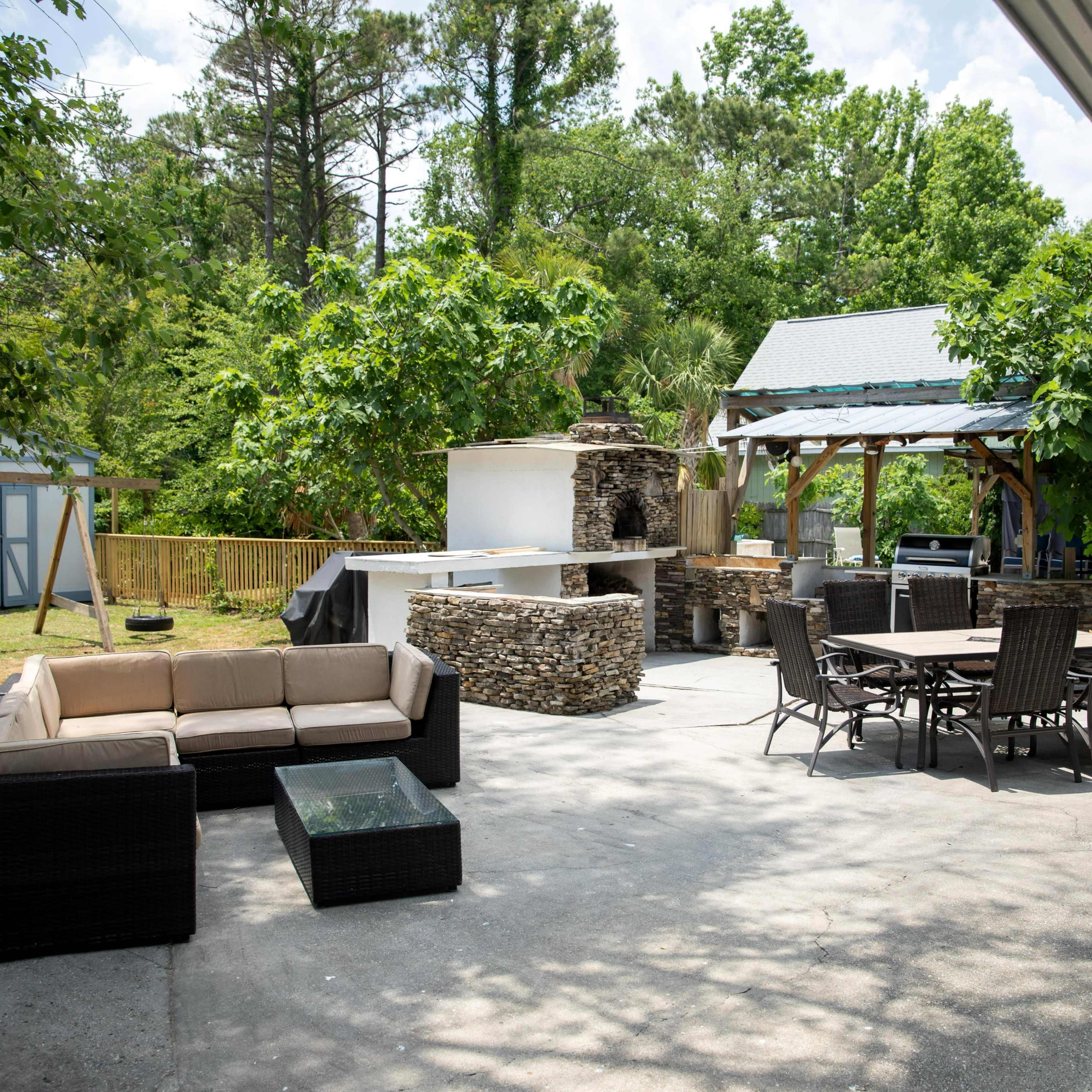 A spacious outdoor patio area featuring a seating arrangement, a dining table, an outdoor kitchen with a stone grill, and various greenery in the background.