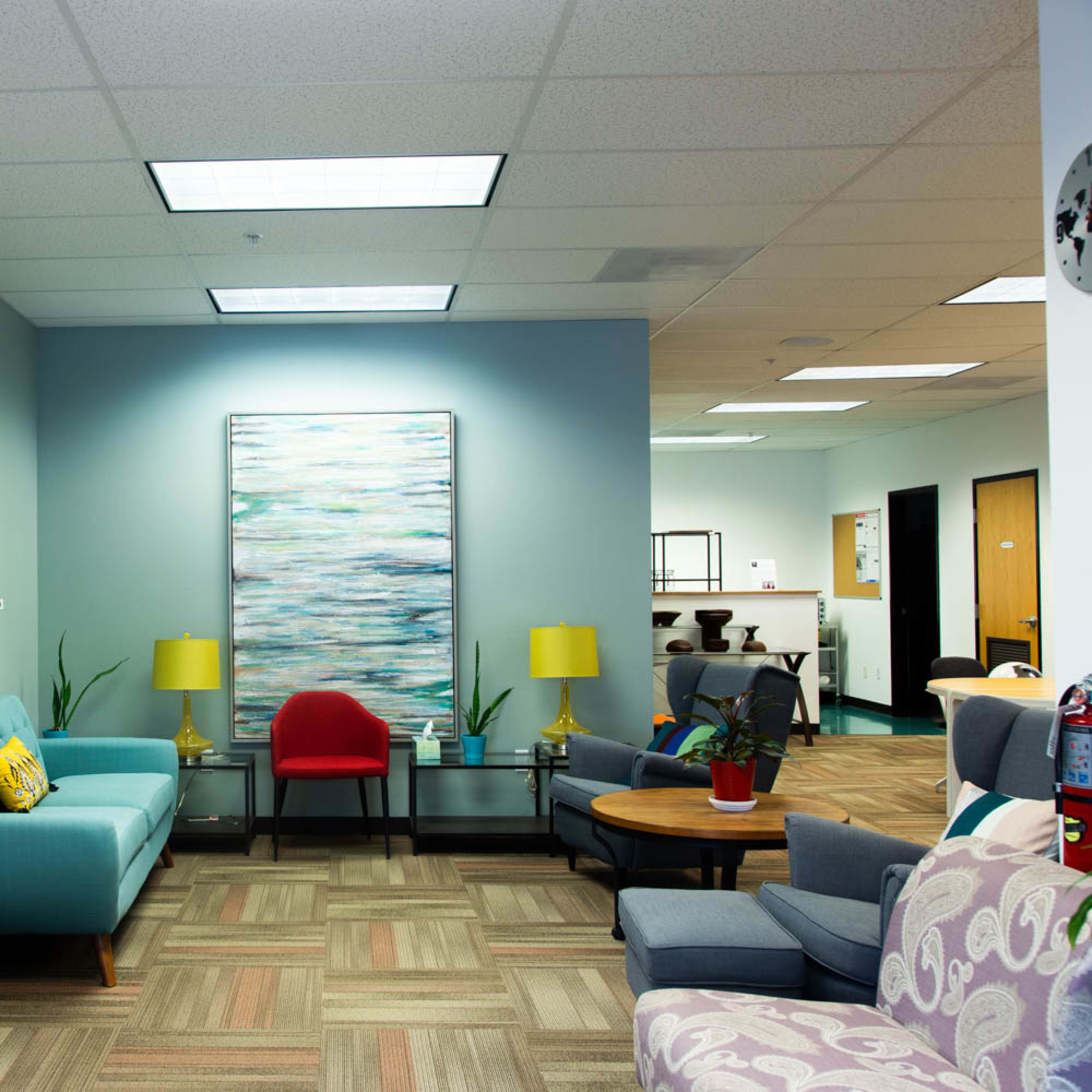 A modern waiting room with a light blue wall, a teal sofa, a red chair, and various artworks, illuminated by ceiling lights.