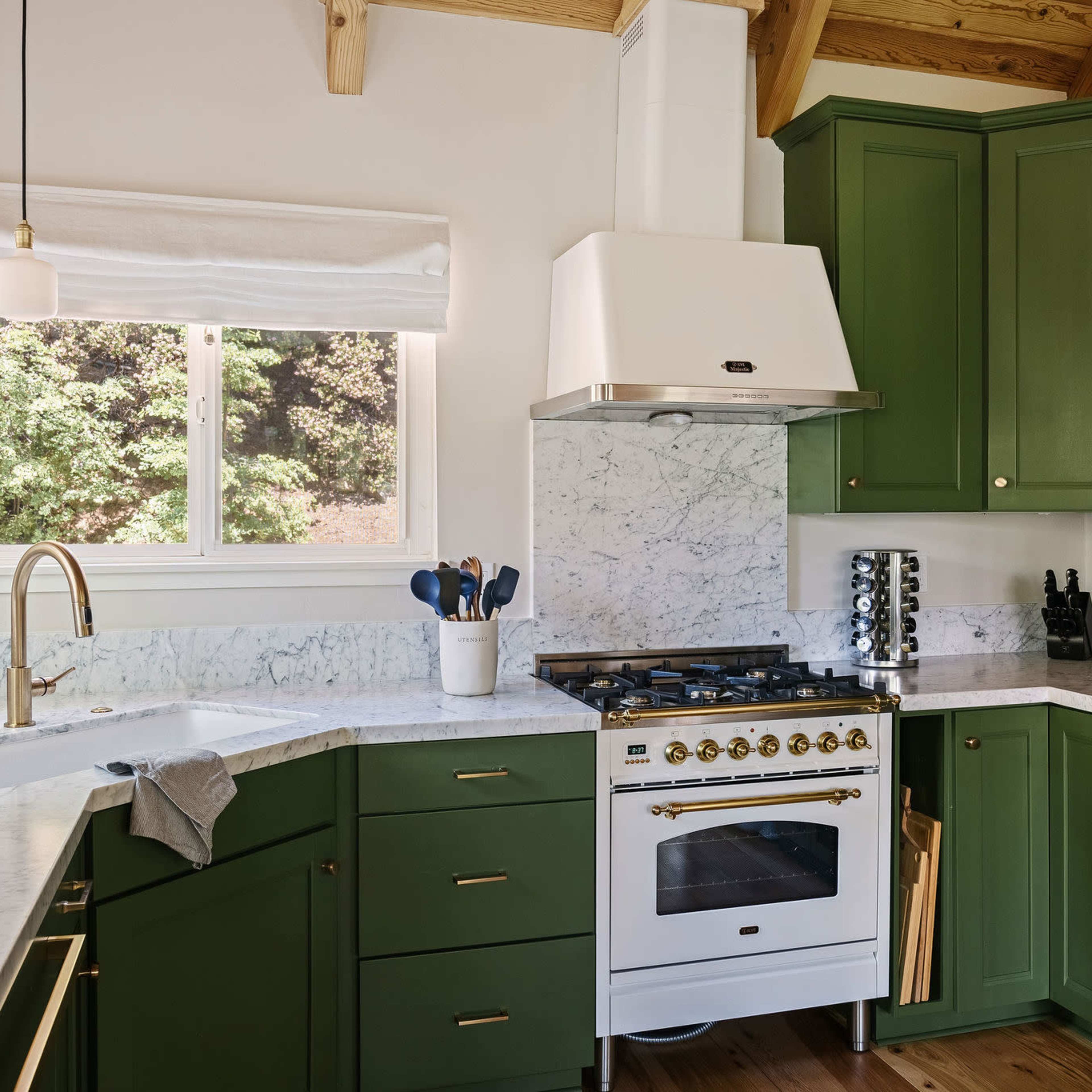 A modern kitchen with green cabinetry, a marble countertop, and a gas stove under a hood, illuminated by natural light from a window.