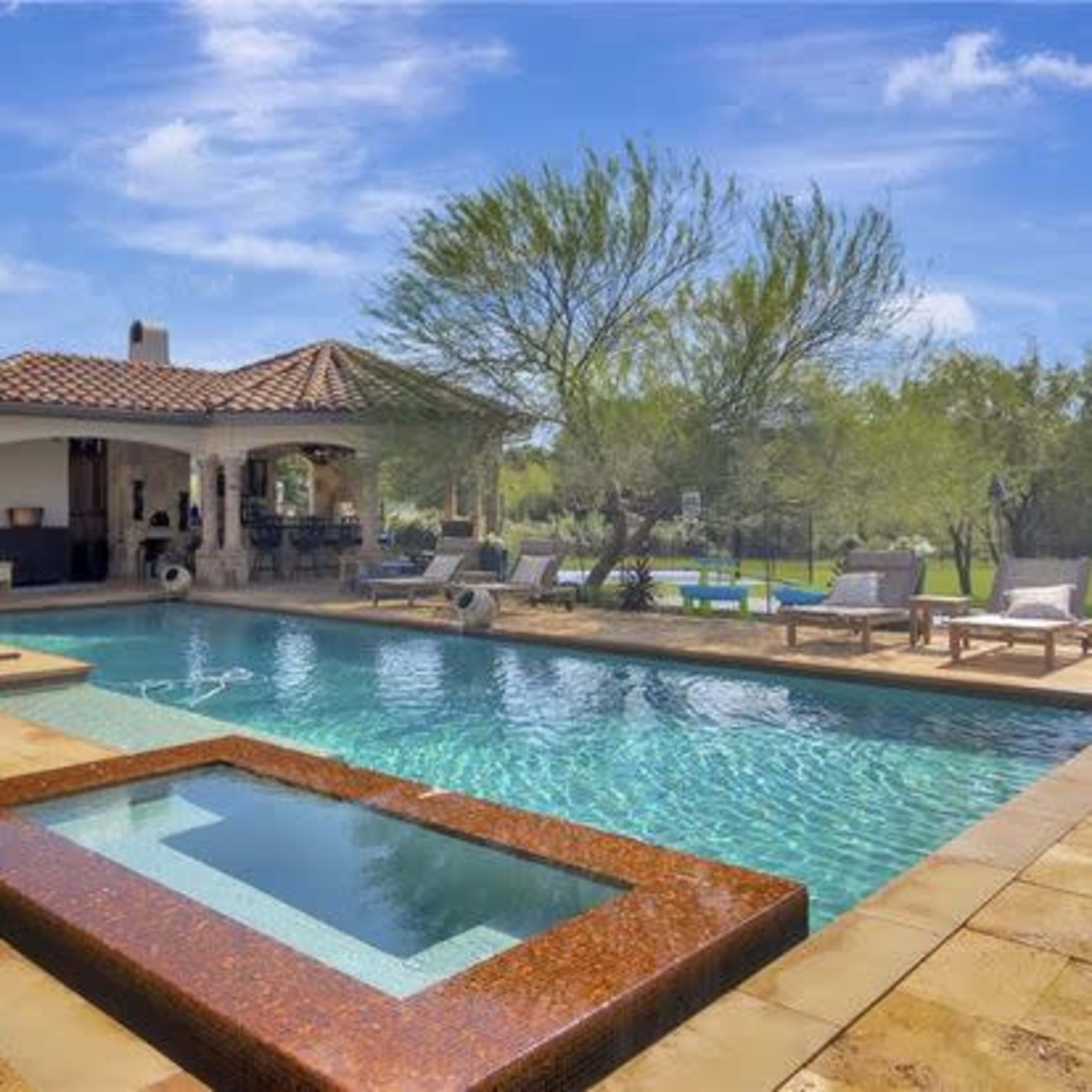 A spacious outdoor pool area featuring a rectangular pool with a spa, surrounded by lounge chairs and a covered patio with dining space.