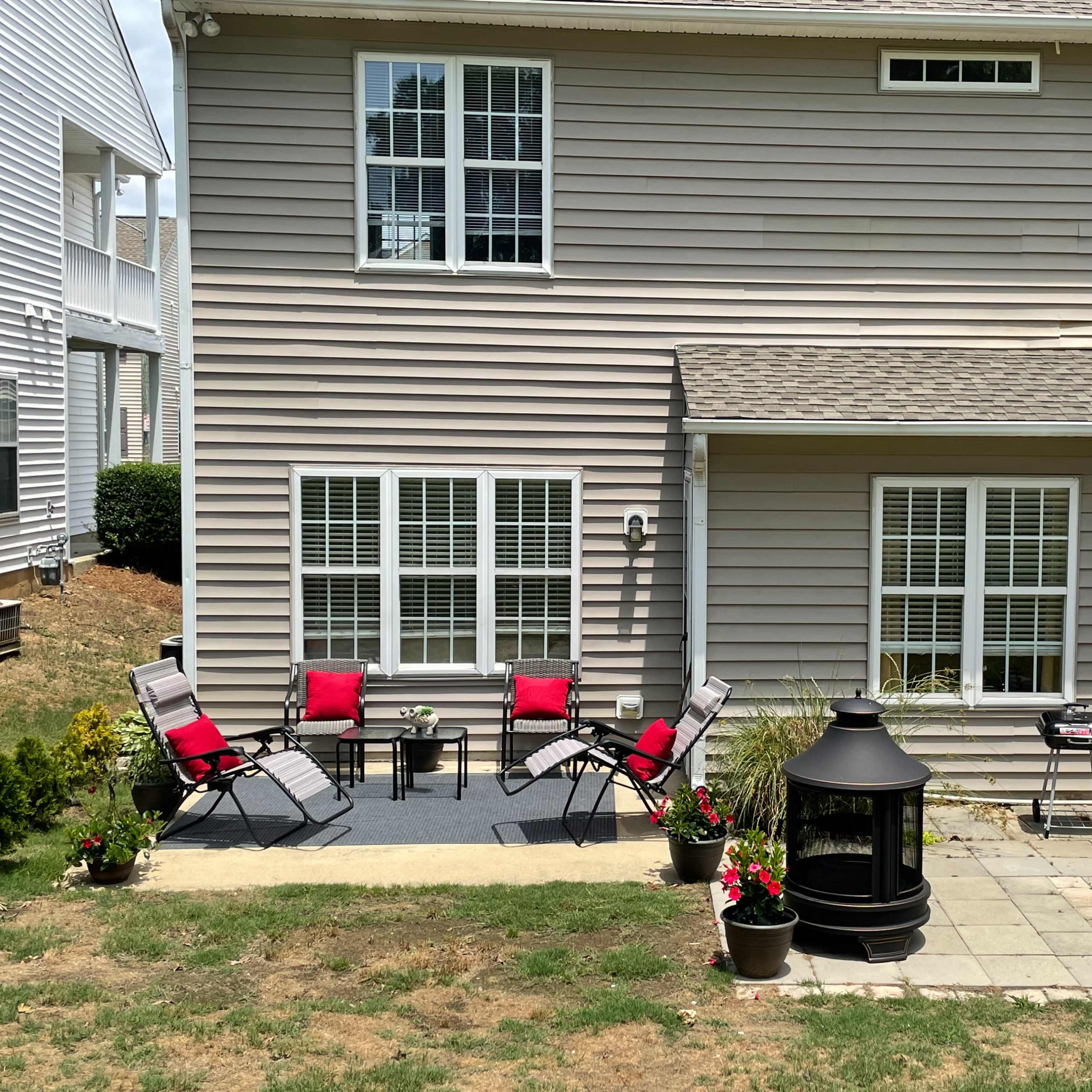 A patio area behind a home with a set of red-cushioned chairs, a small table, a grill, and planters with flowers.