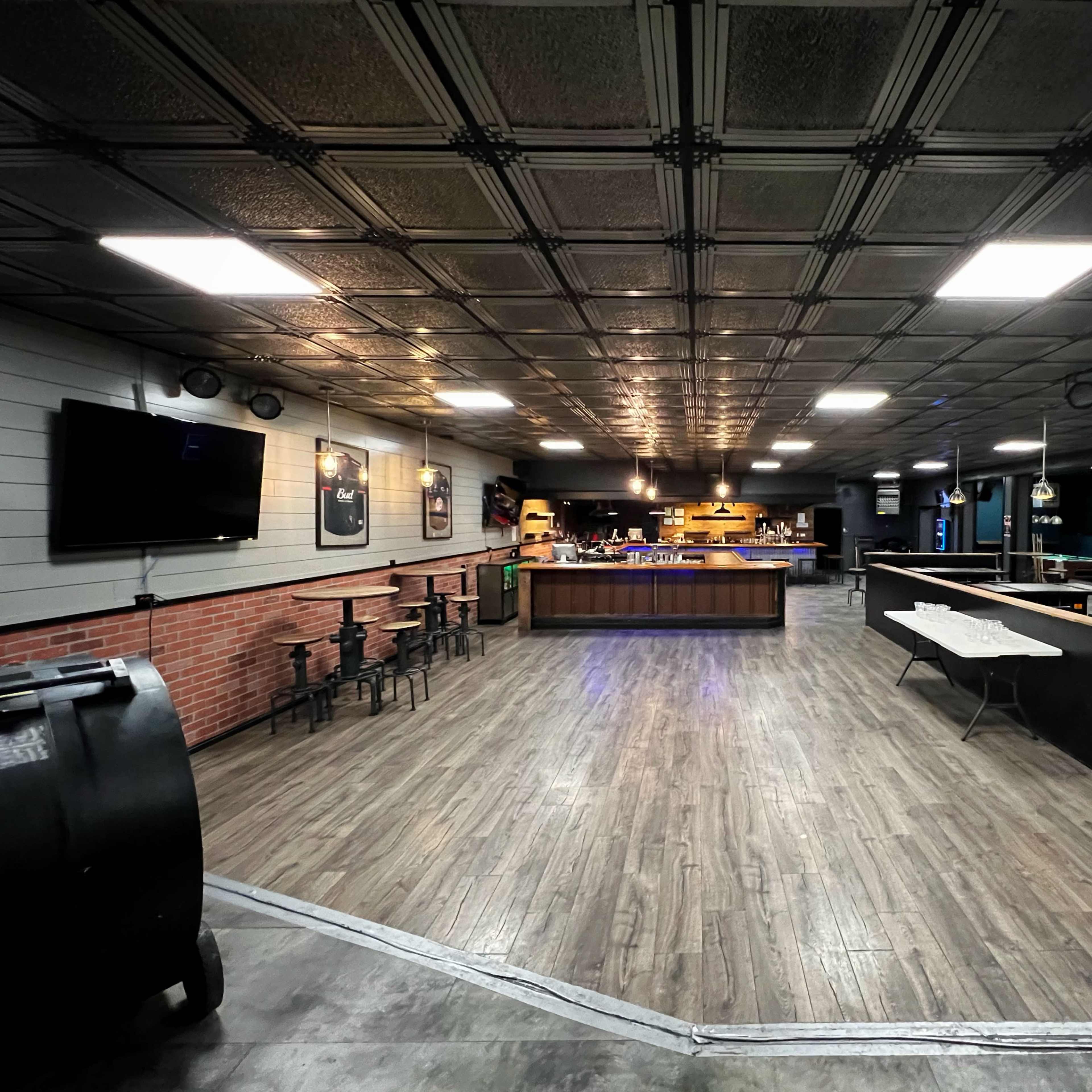 A spacious, dimly-lit interior of a bar or entertainment venue with a wooden bar counter, seating areas, and a patterned ceiling.