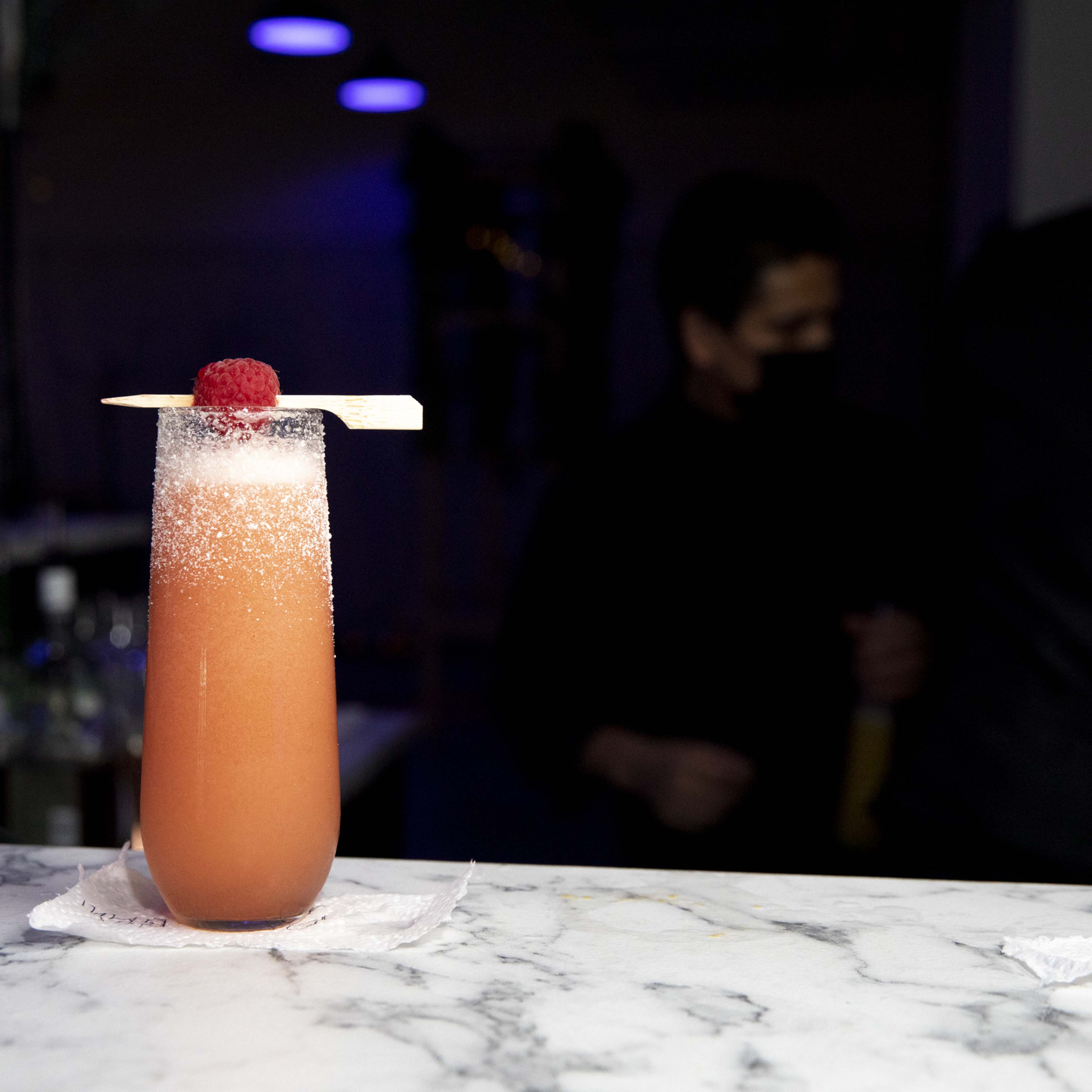 A cocktail garnished with a raspberry and served in a tall glass sits on a marble bar.