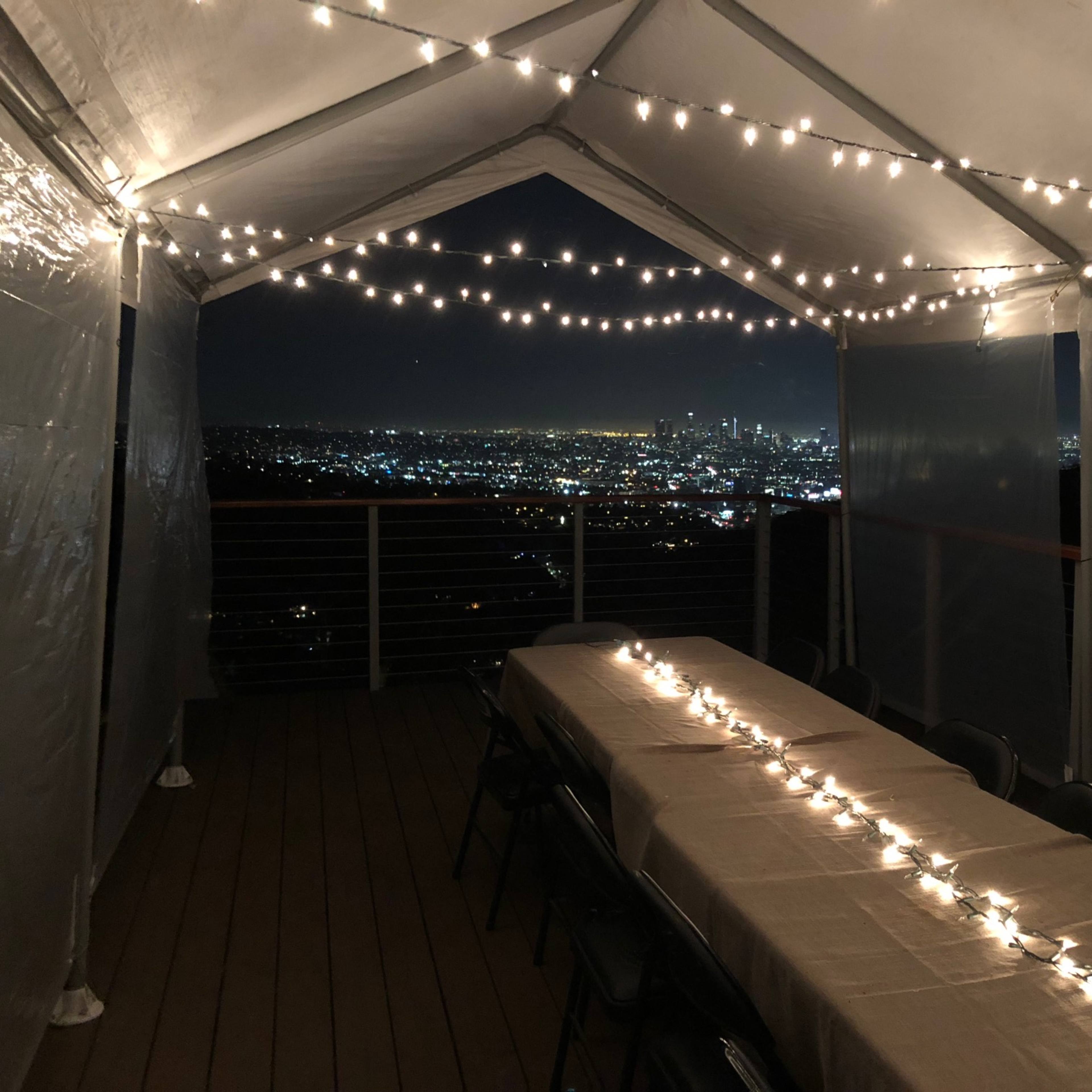 A long table is set under a tent adorned with string lights, overlooking a city skyline at night.