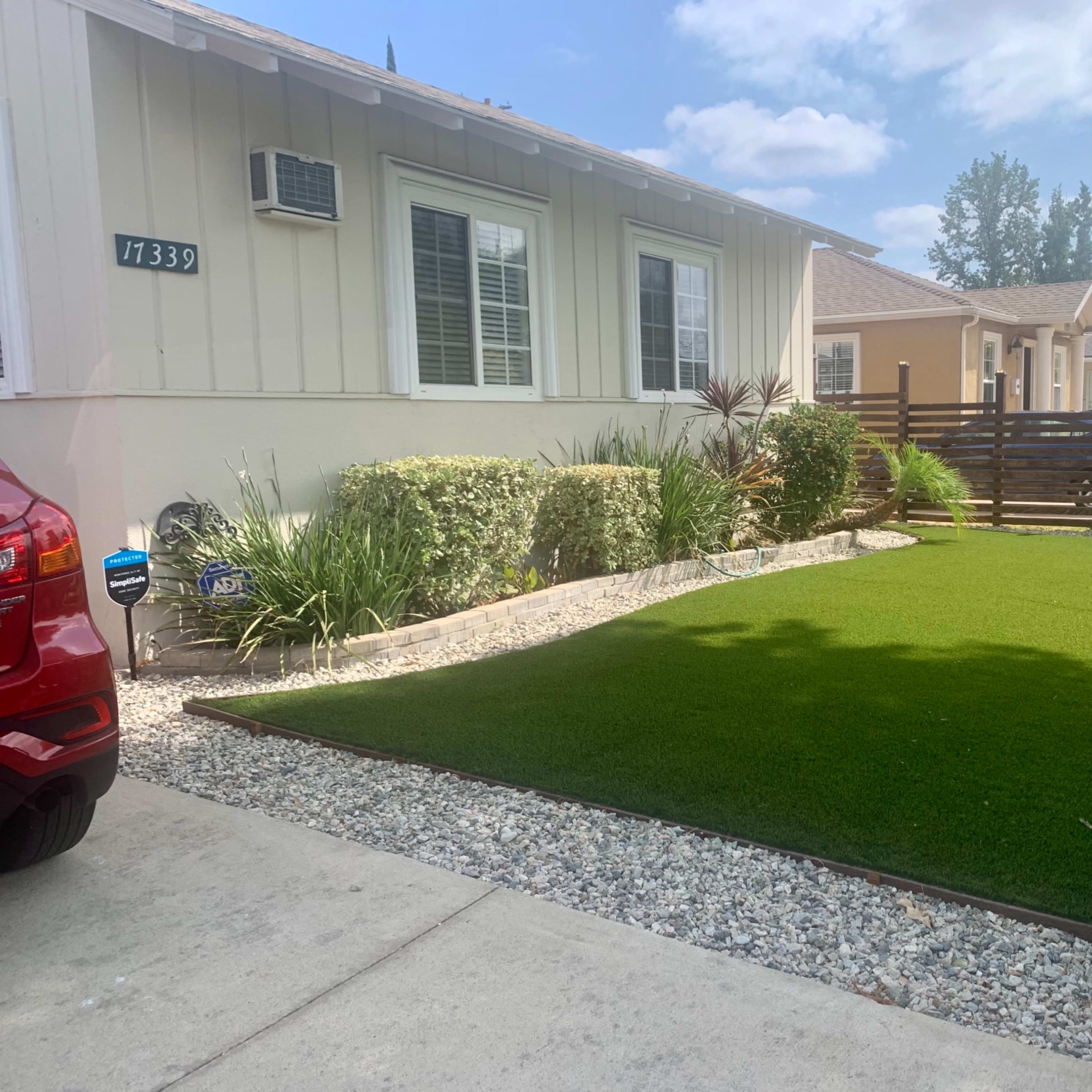 A residential front yard with artificial grass bordered by gravel and low shrubs, alongside a red car parked on the driveway.