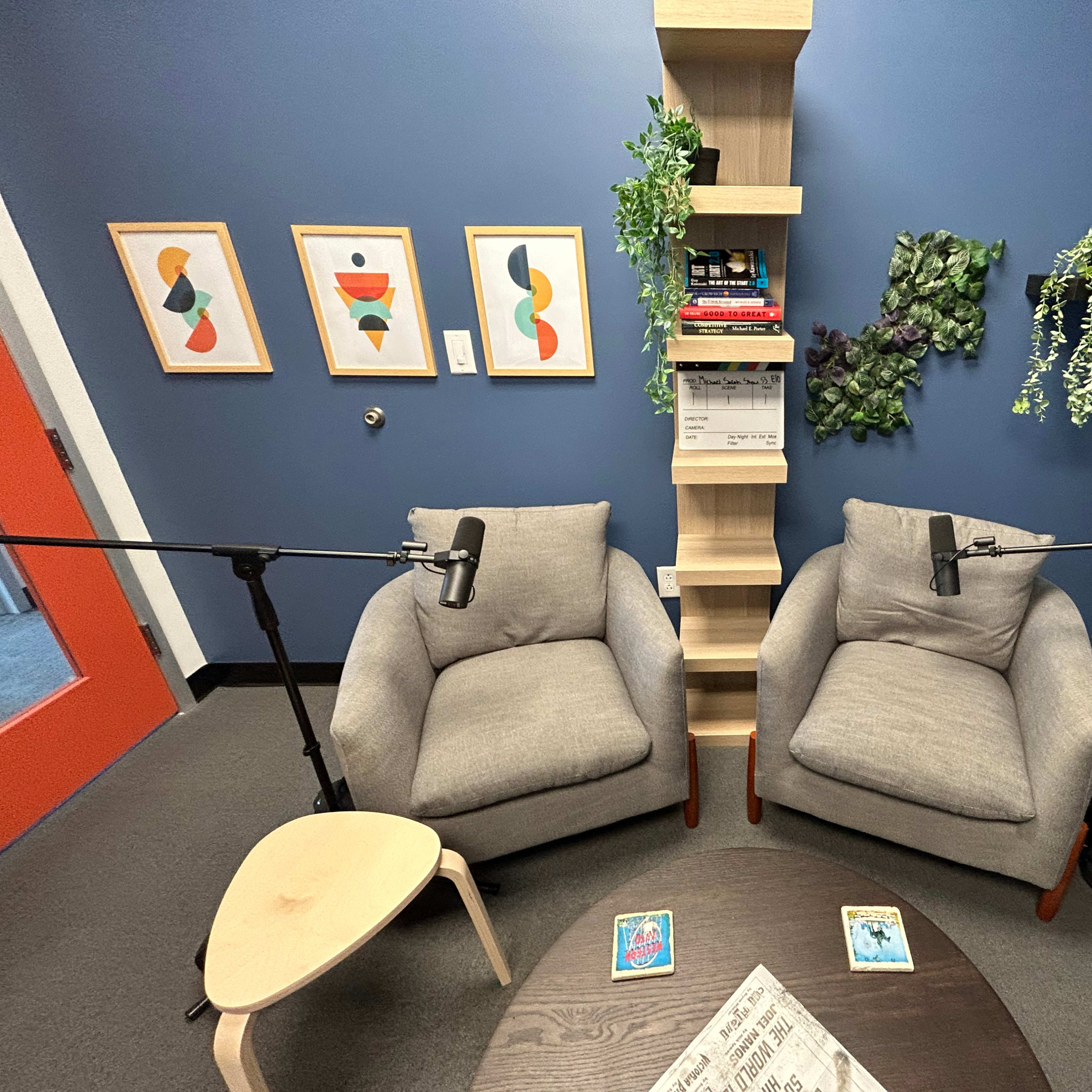 A small recording studio setup with two gray chairs, a round table, and microphones positioned between them, surrounded by wall art and shelves containing books and plants.