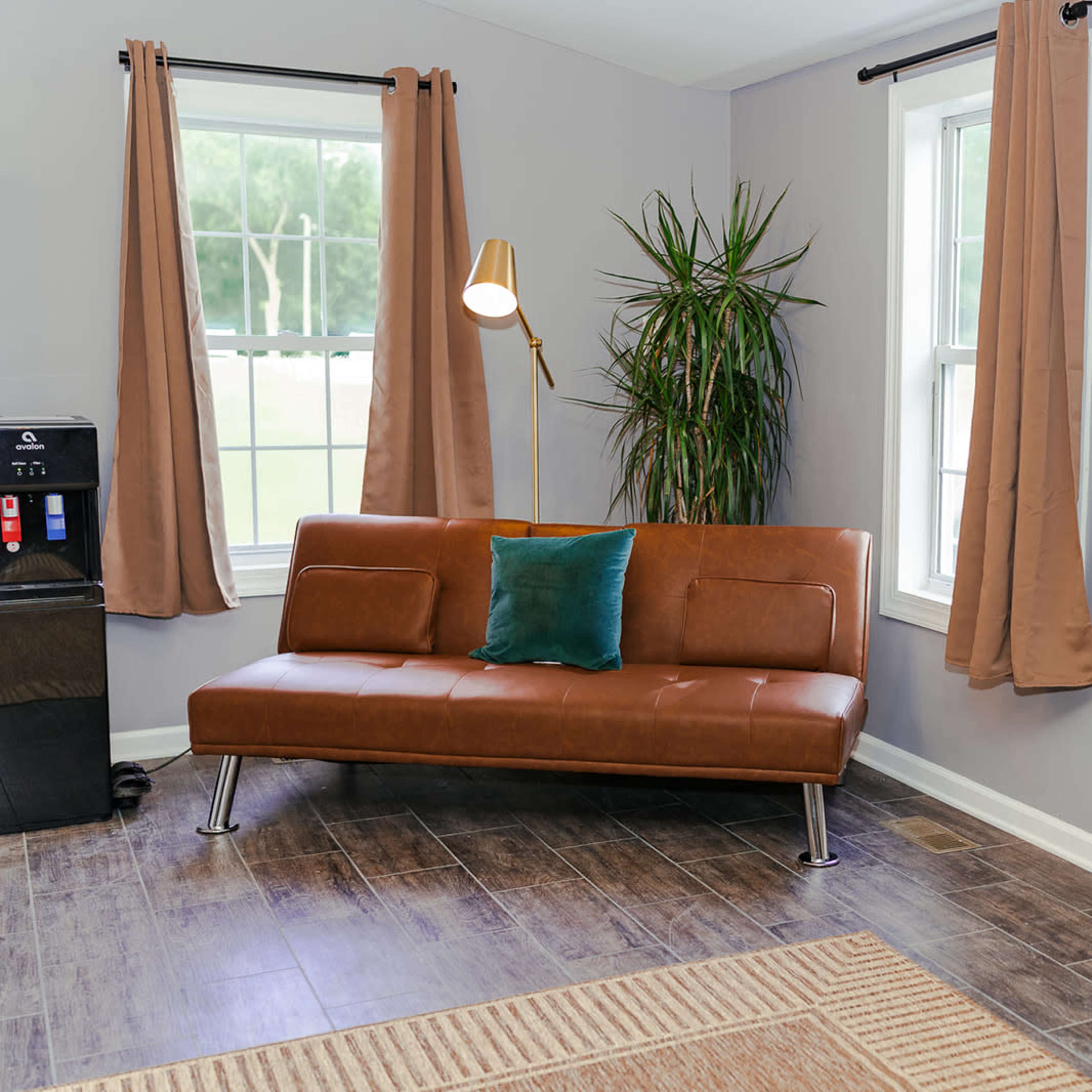 A living room with a brown sofa, a potted plant, a dartboard on the wall, and a vintage-style radio cabinet beside it.