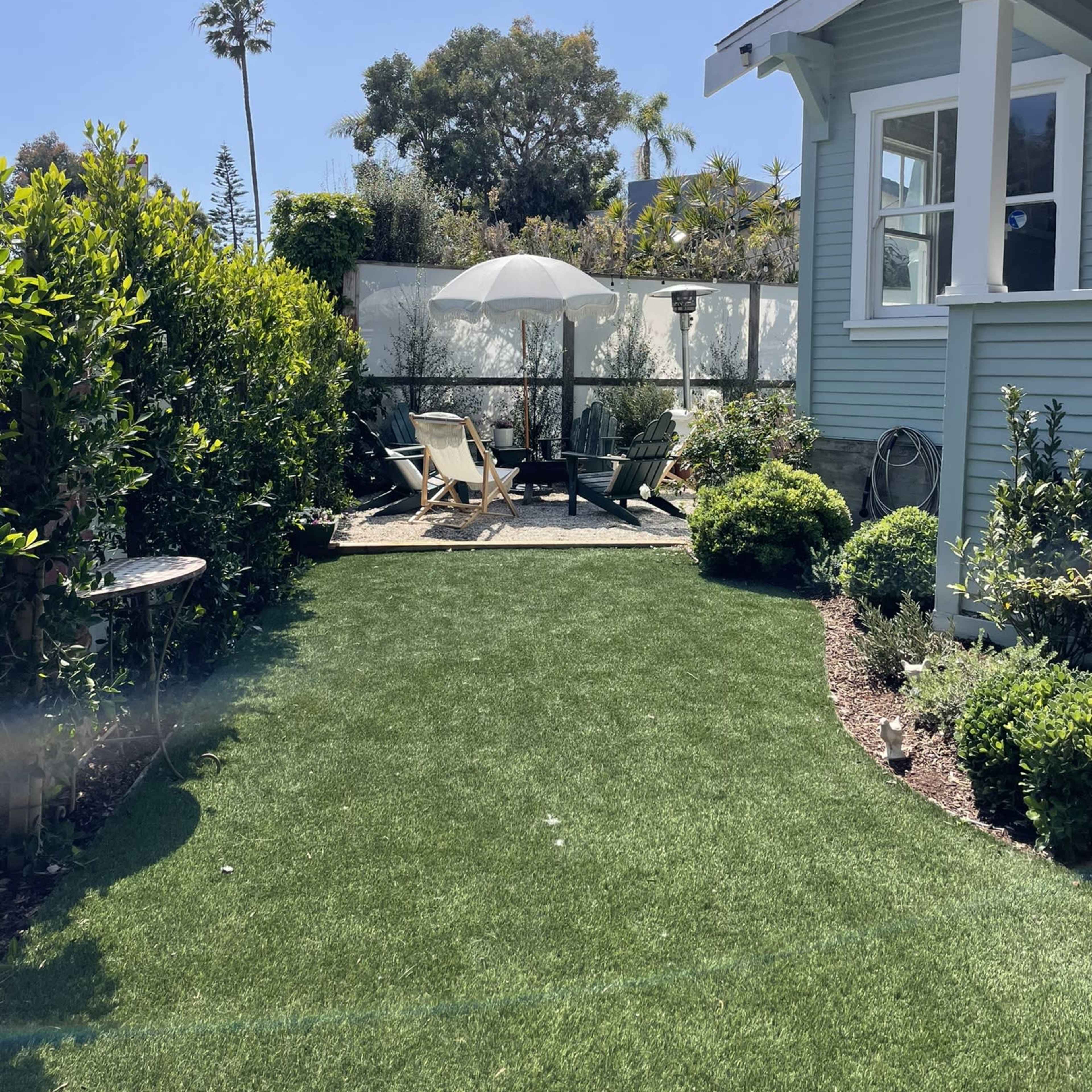 A sunny backyard garden with artificial grass, featuring a lounge chair, an umbrella, and neatly trimmed hedges.