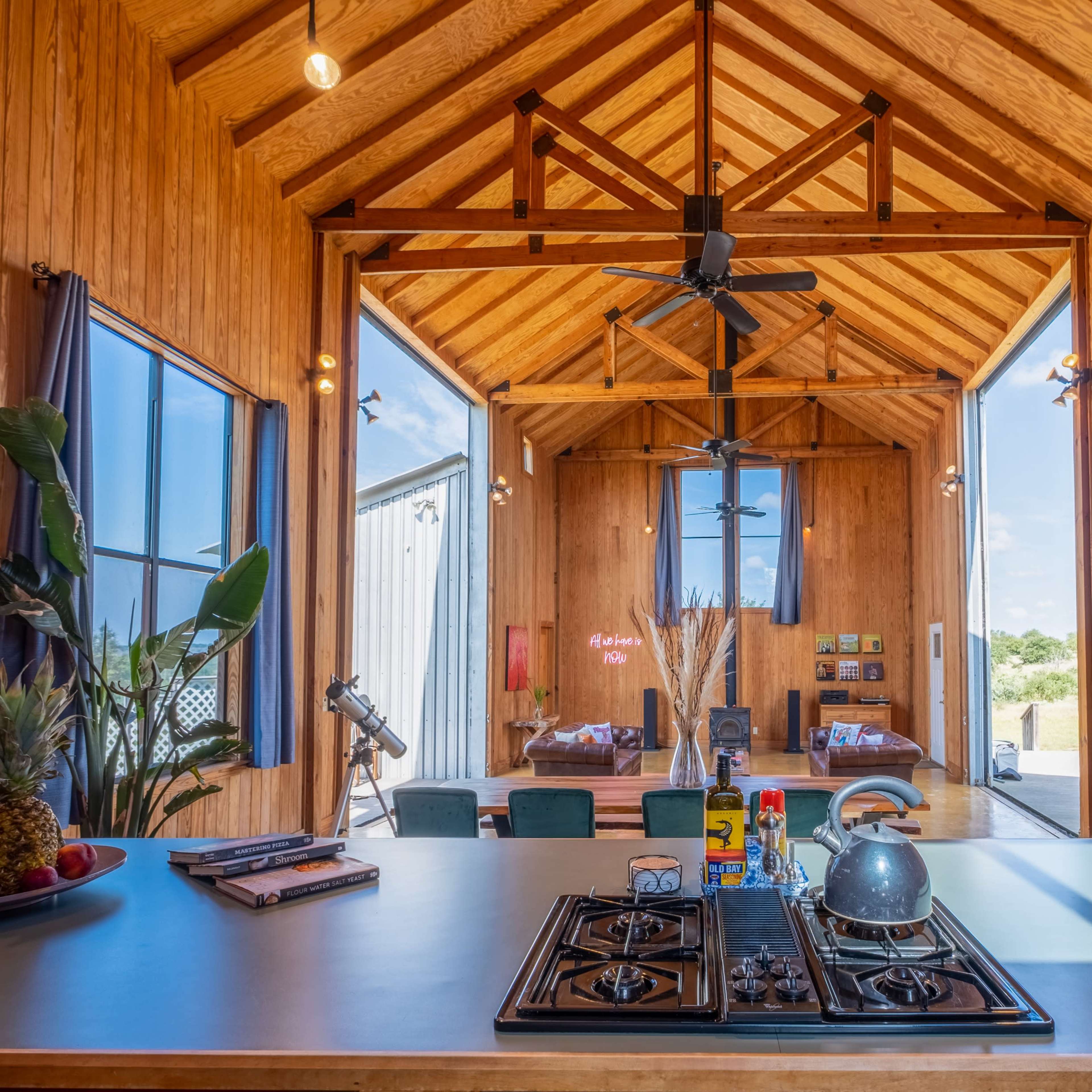 A wooden kitchen with a central island, featuring a gas stove, a dining area, and large windows allowing natural light to enter.