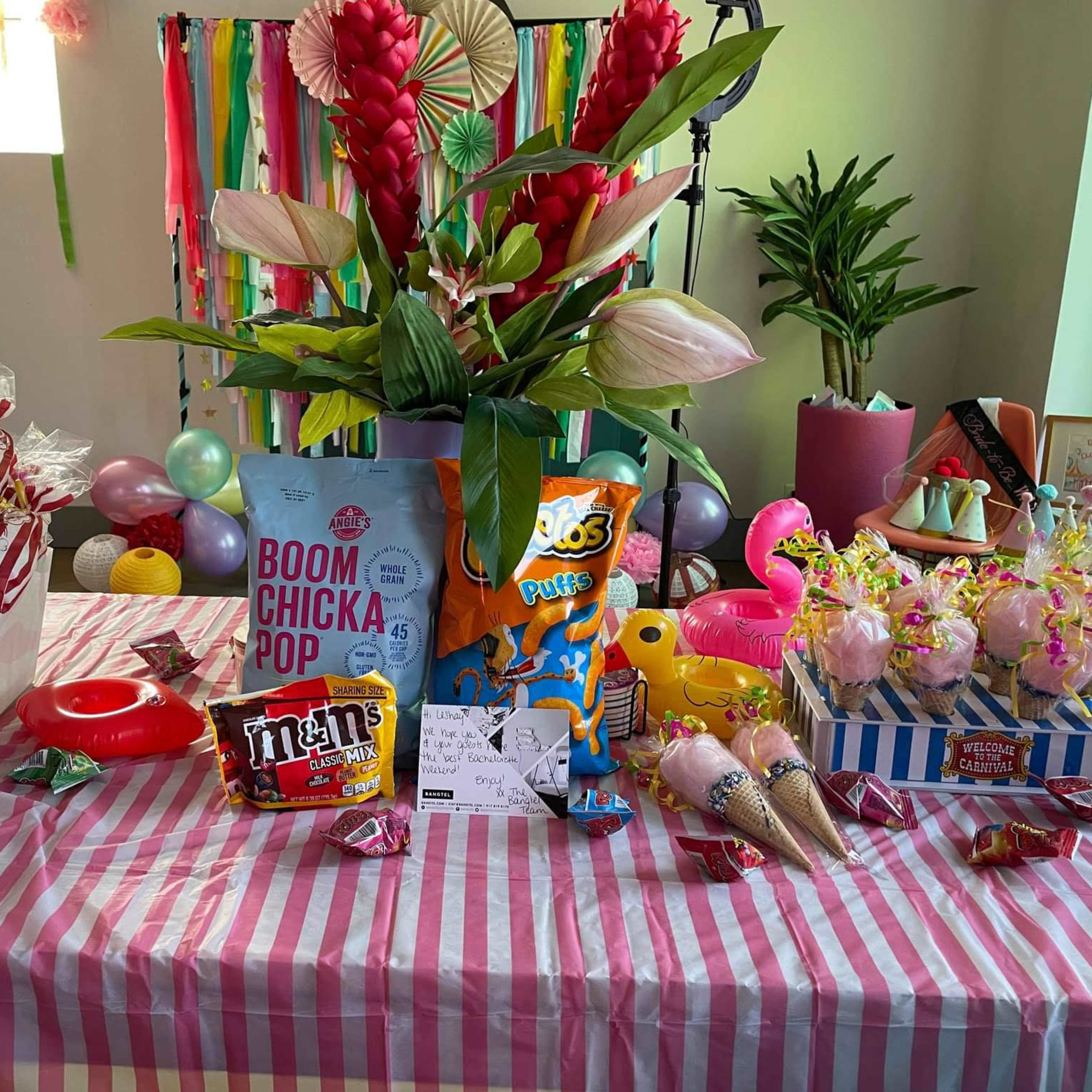 A colorful party table is decorated with snacks and treats, including popcorn, candies, and floral arrangements.