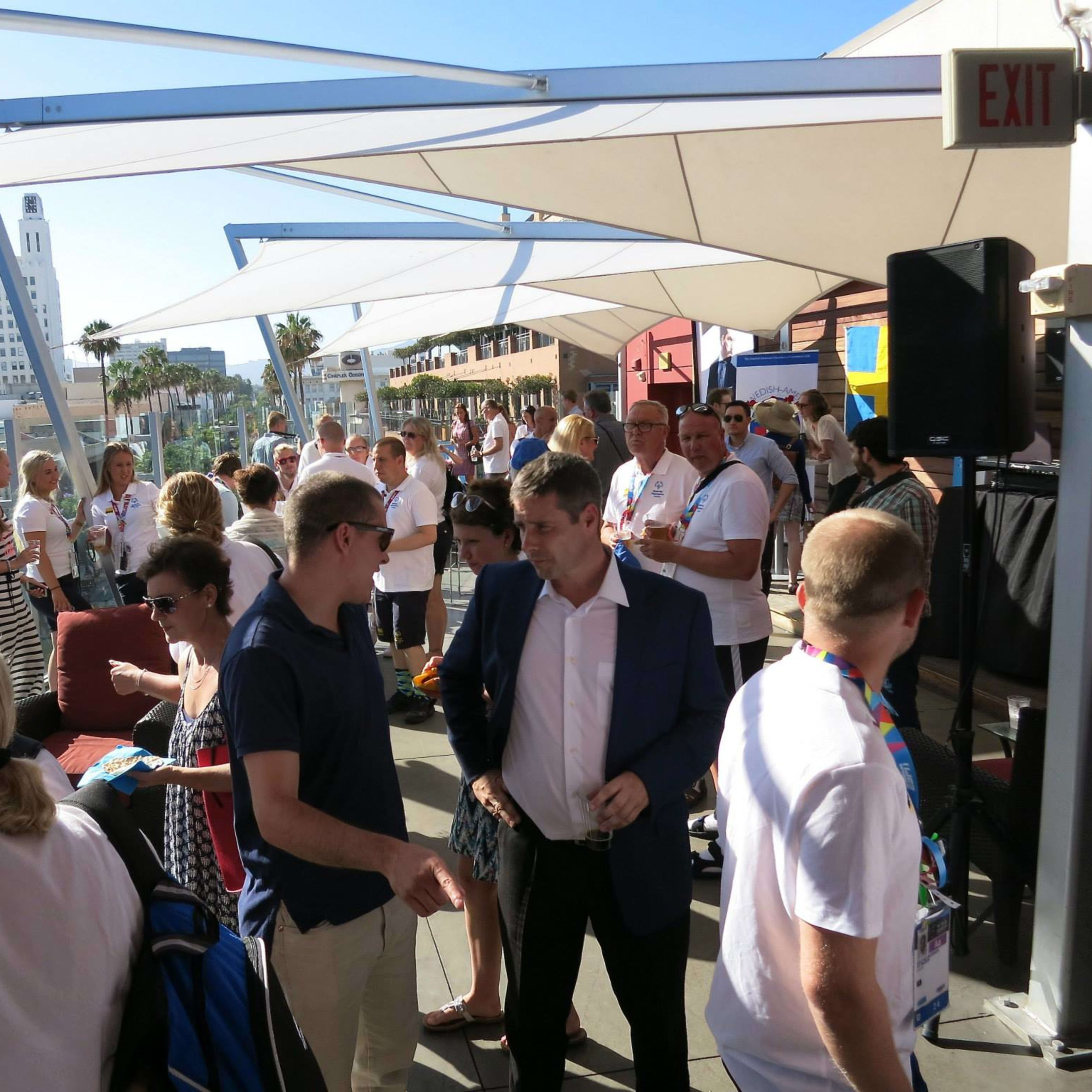 A crowded rooftop event features attendees mingling under large shaded canopies with a city skyline in the background.