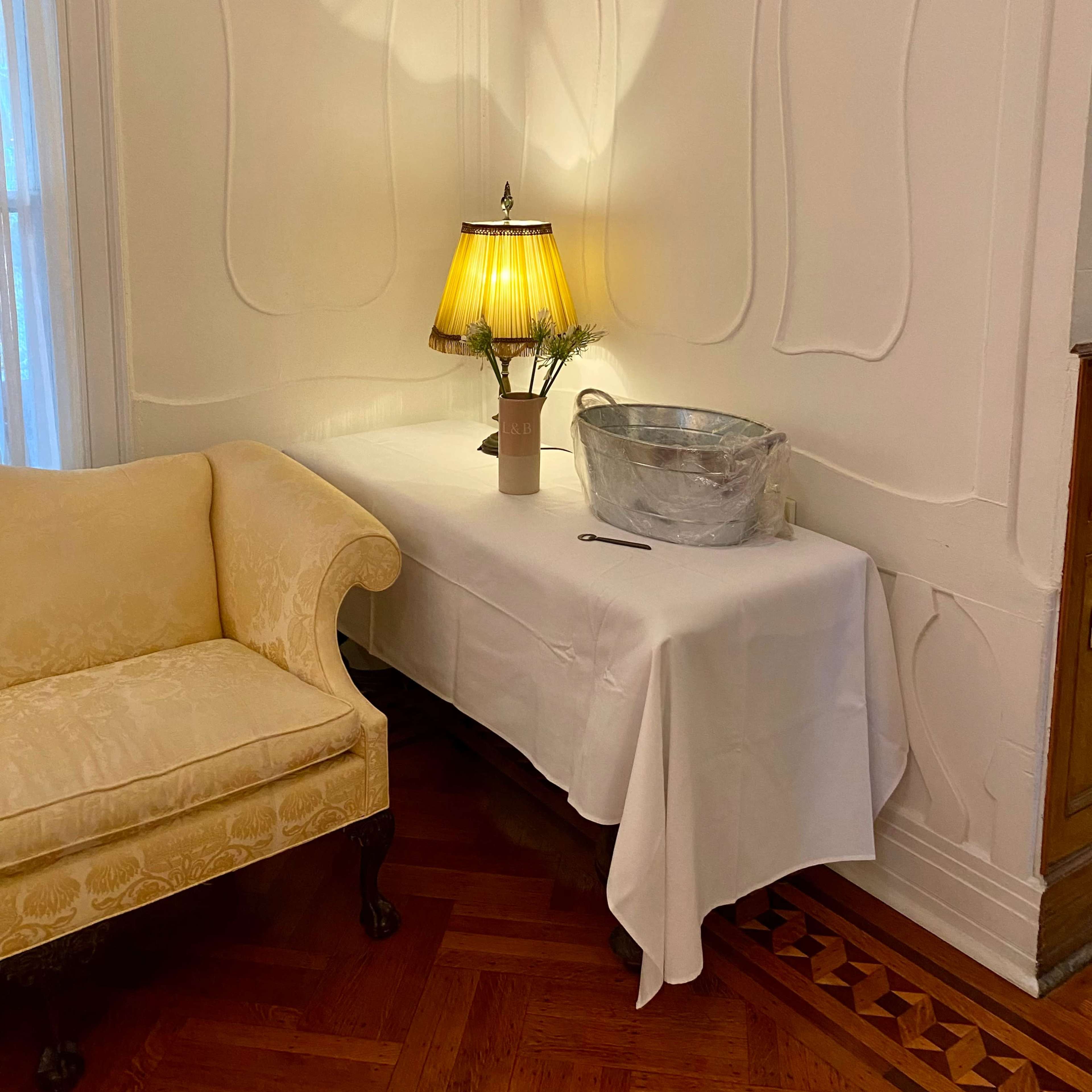 A decorative corner of a room features a yellow lamp on a table draped with a white cloth, alongside a metallic bowl.