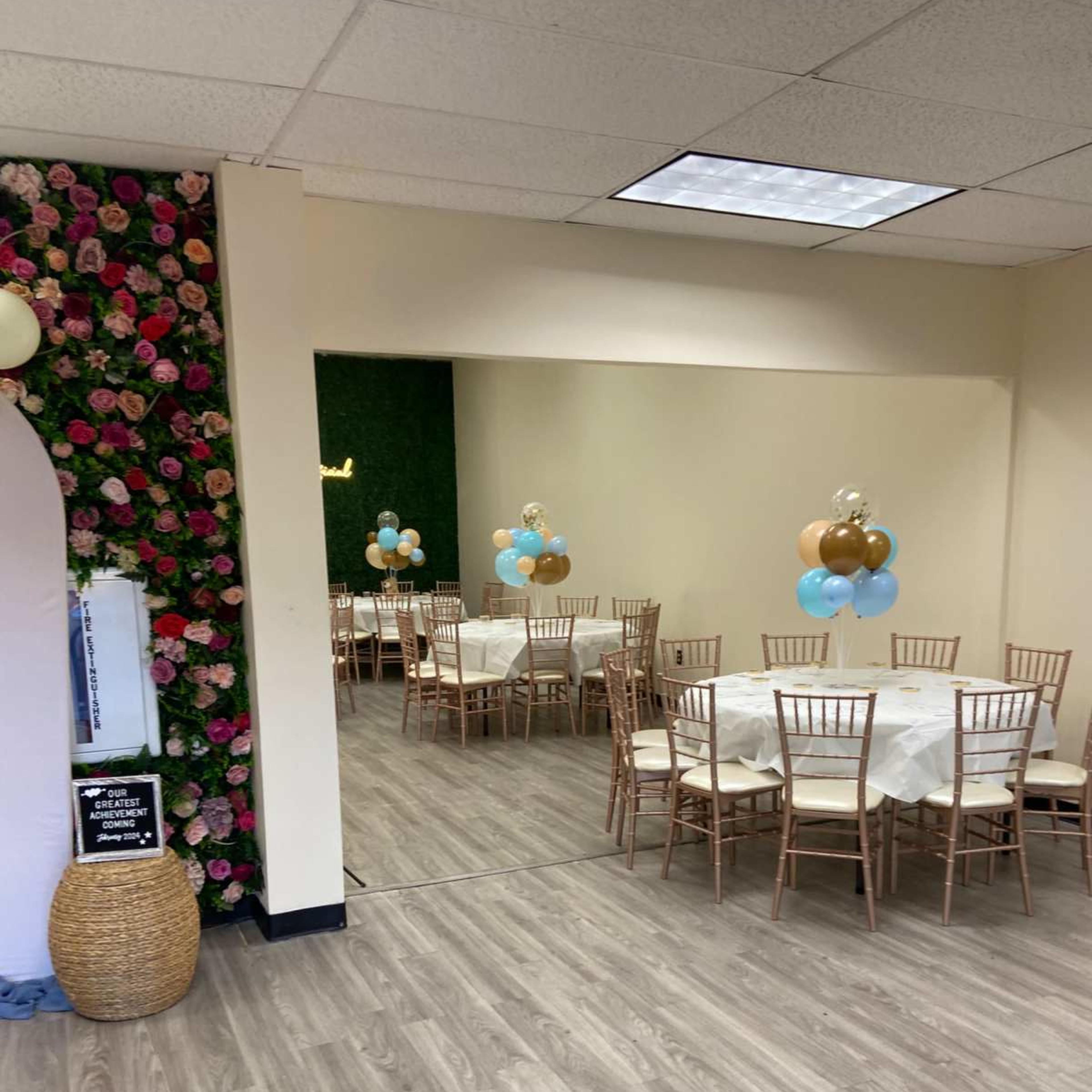 A decorated event space with multiple tables set with white tablecloths and chairs, along with balloon arrangements in blue and gold.