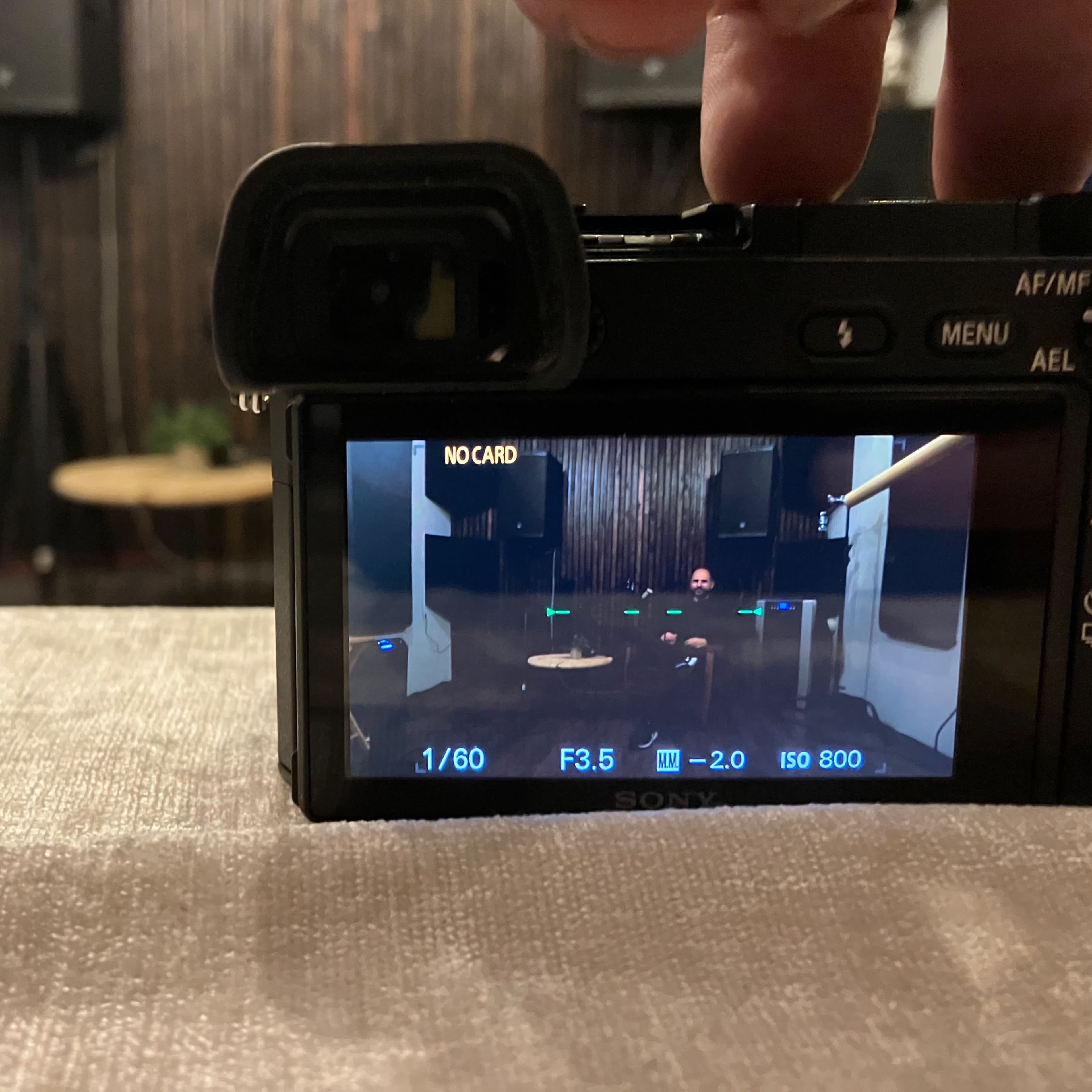 A camera on a table captures a view of a person seated in a dimly lit room with wooden paneling and sound equipment.