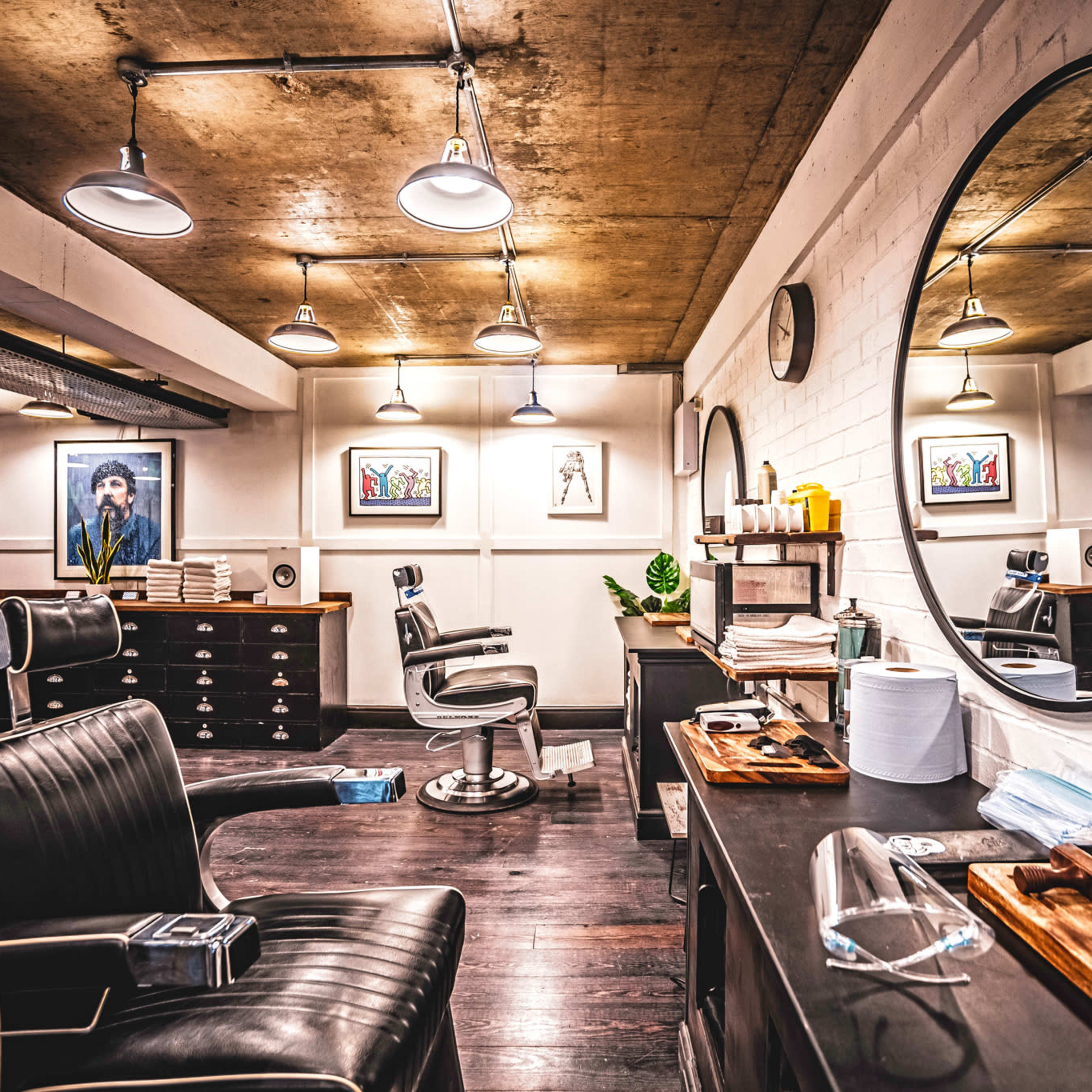 A modern barbershop interior featuring two barber chairs, a large circular mirror, and framed art on the walls.
