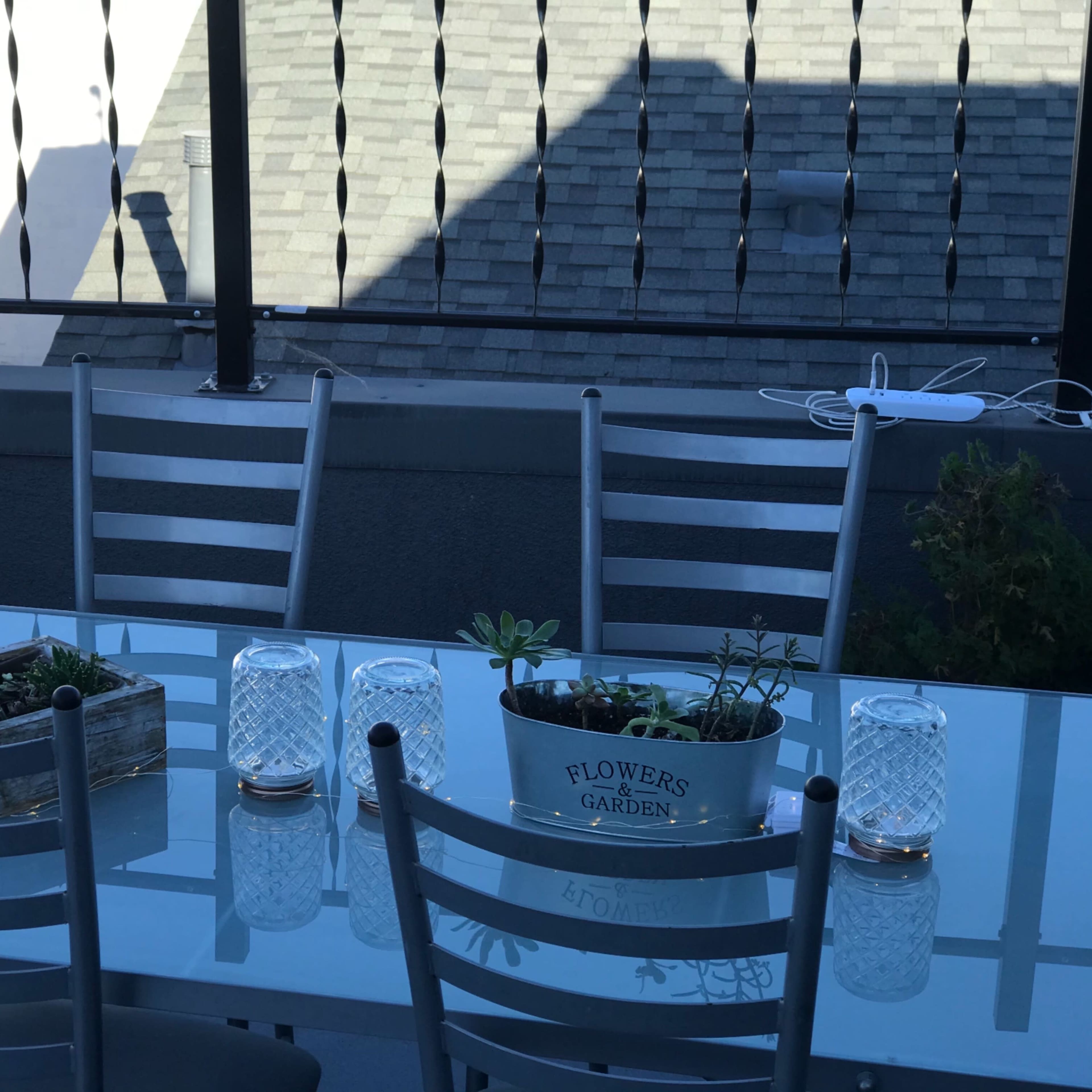 A glass-topped outdoor dining table with metal chairs is set on a balcony, featuring a planter with succulents and glass candle holders.