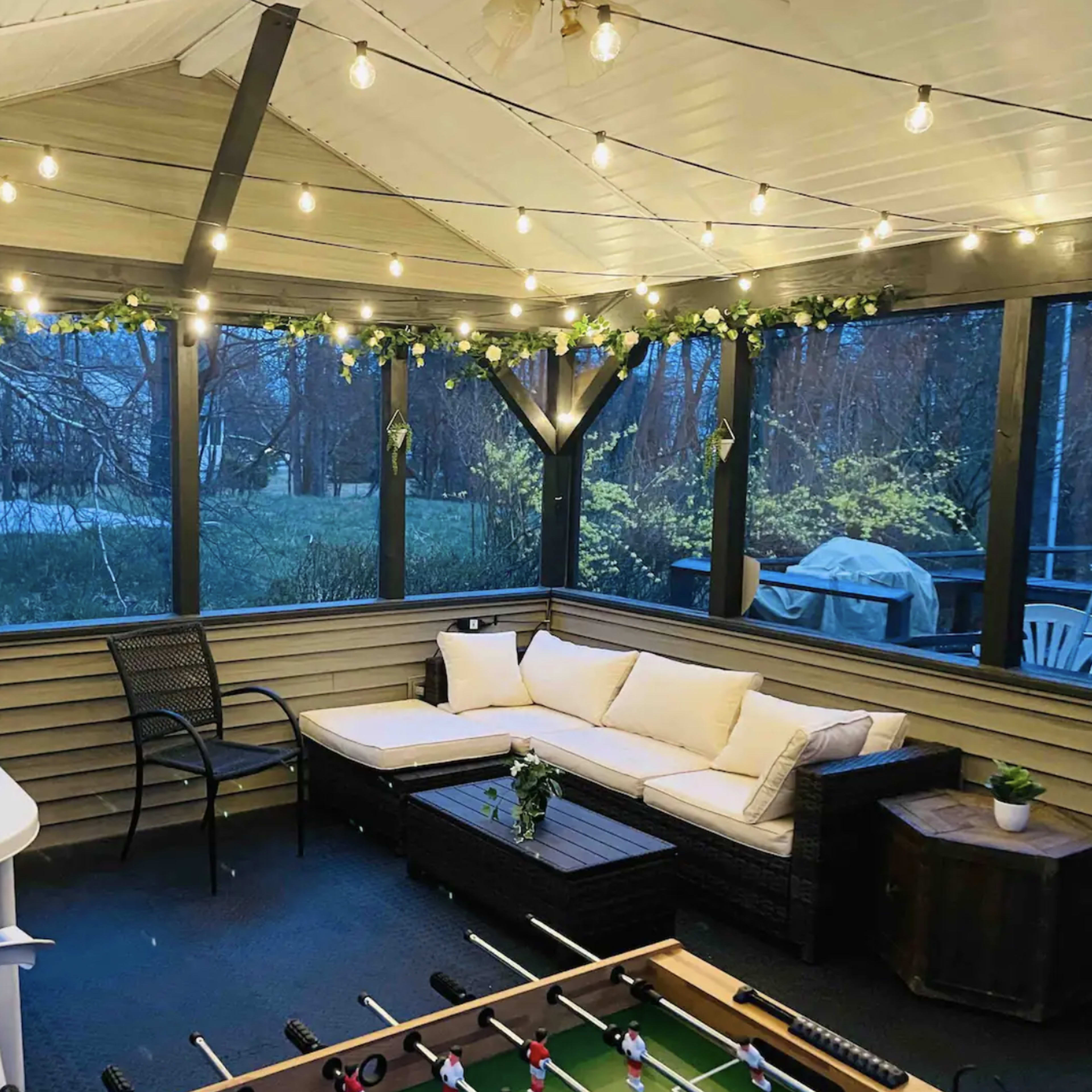 A screened-in porch featuring comfortable seating, a foosball table, and decorative string lights.