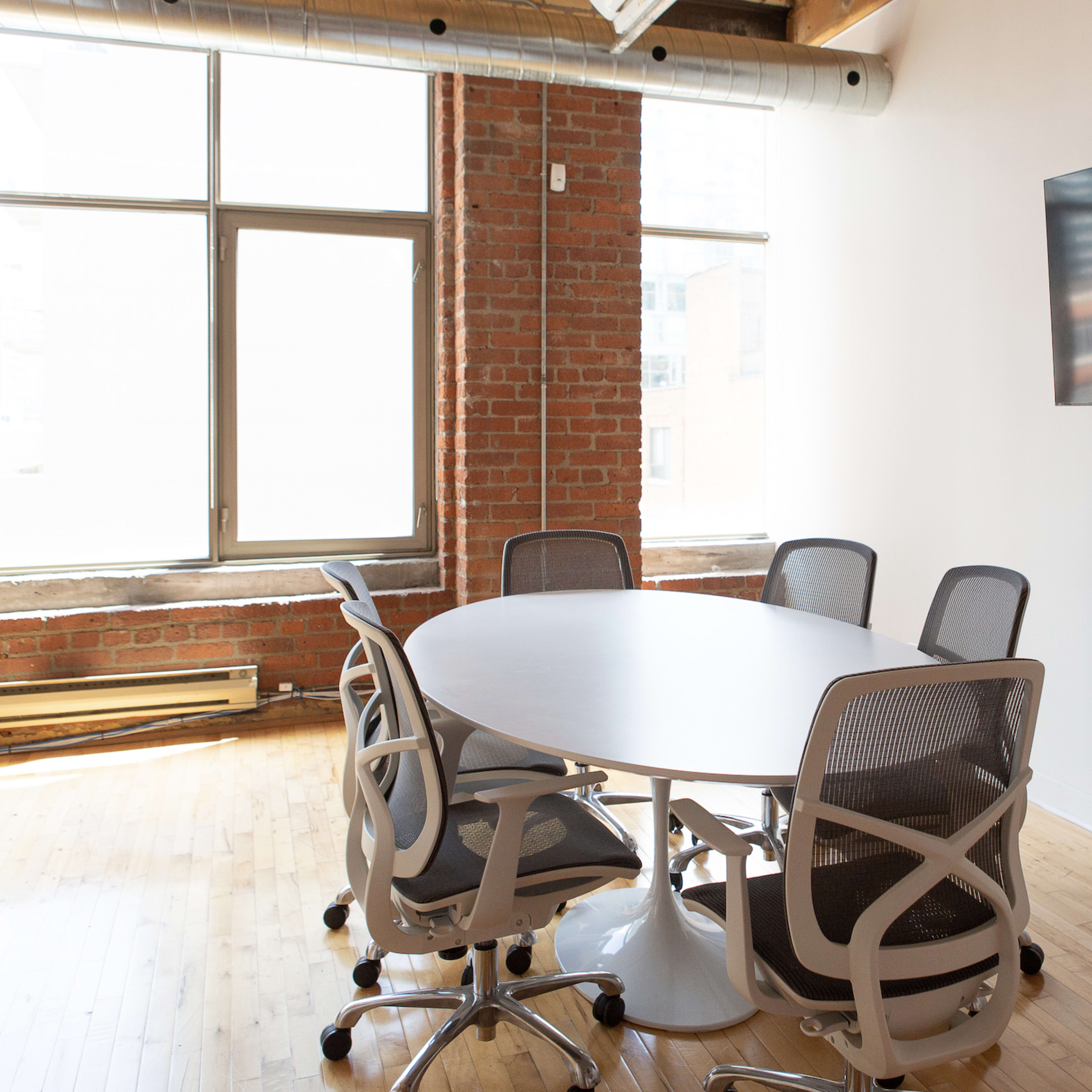 A modern conference room featuring a round table surrounded by five mesh-backed chairs and a flat-screen TV mounted on the wall.