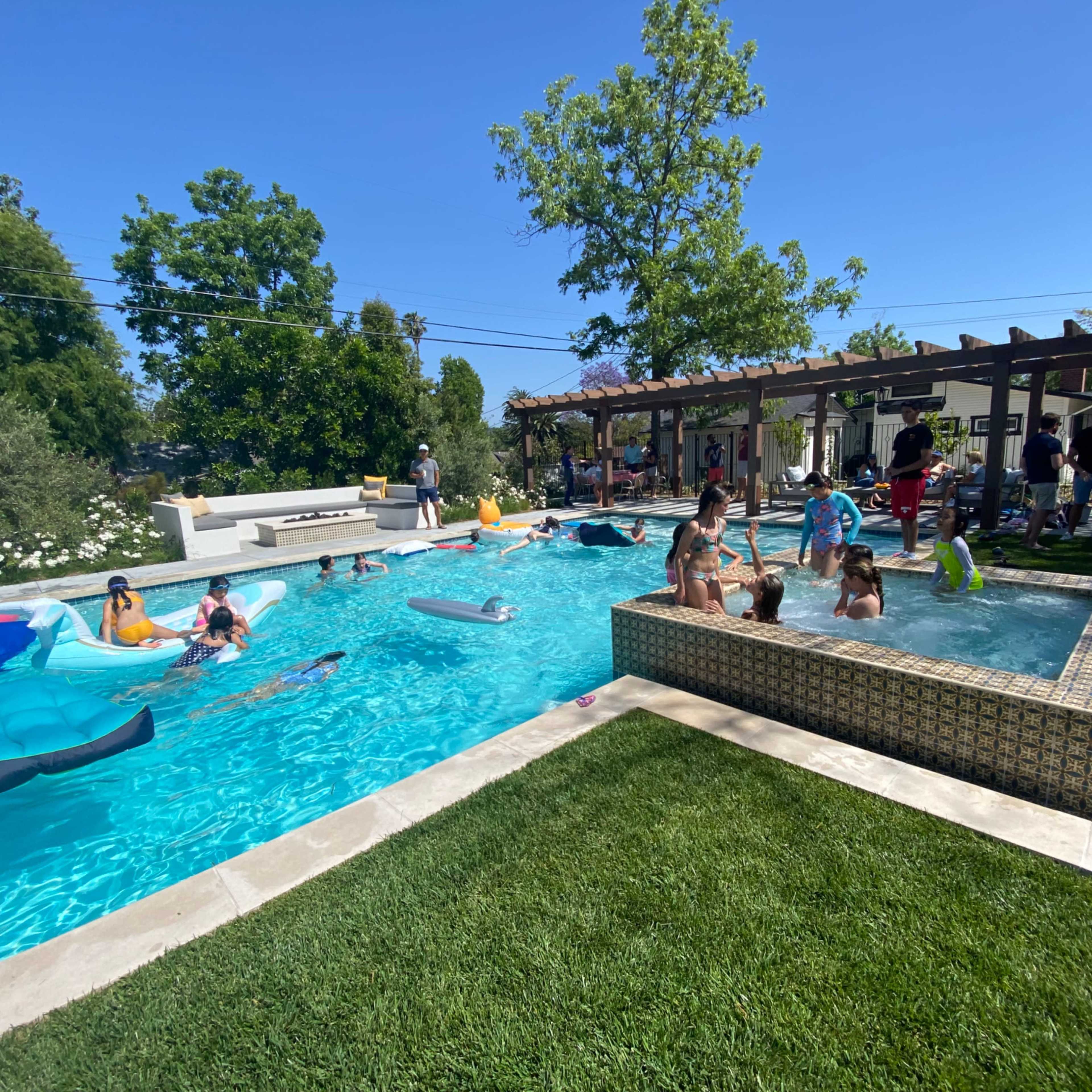 The scene shows a large swimming pool surrounded by people enjoying the water, with colorful floats and a grassy area nearby.