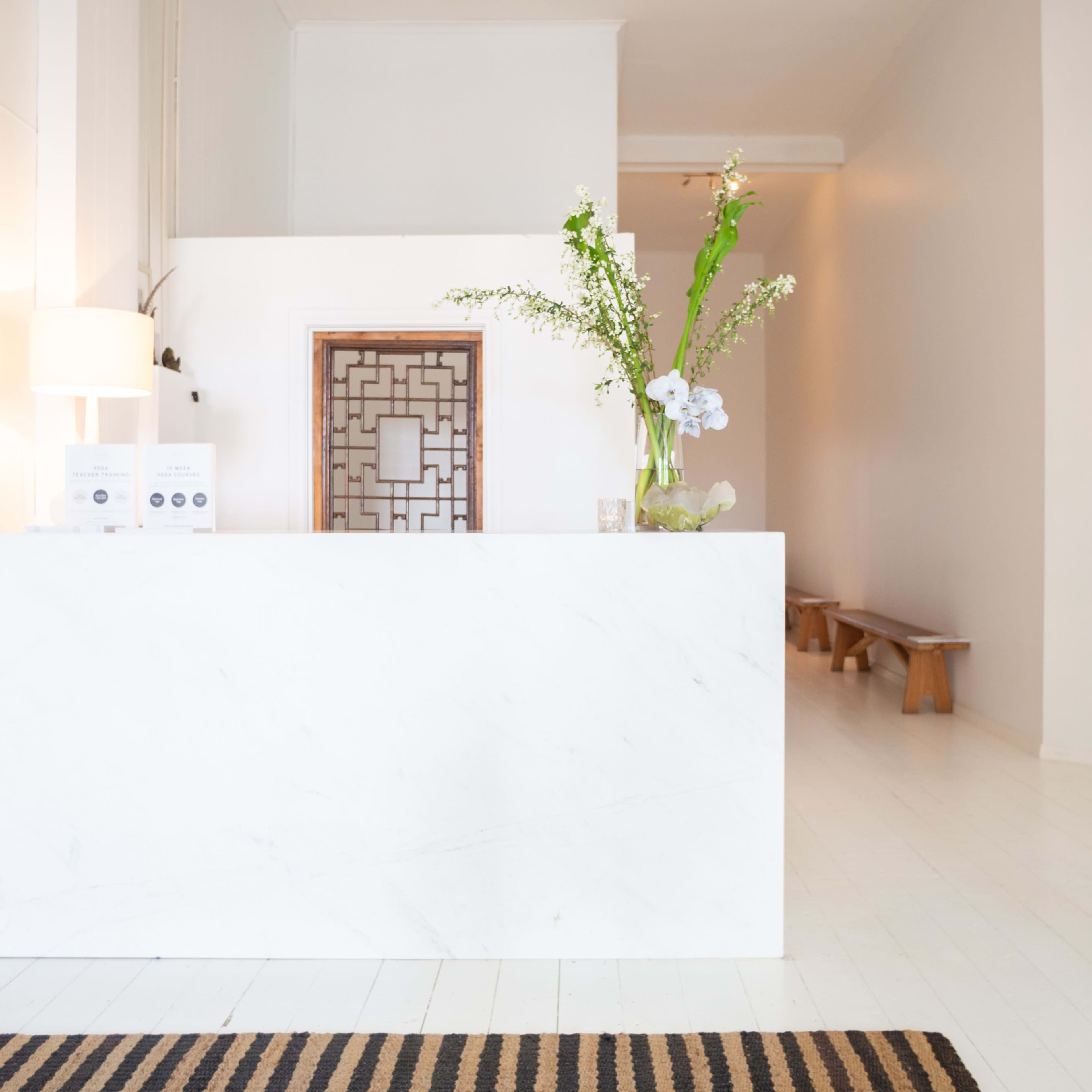 A modern reception area with a white marble front desk, a decorative mirror, and a minimalistic waiting area featuring a wooden bench.