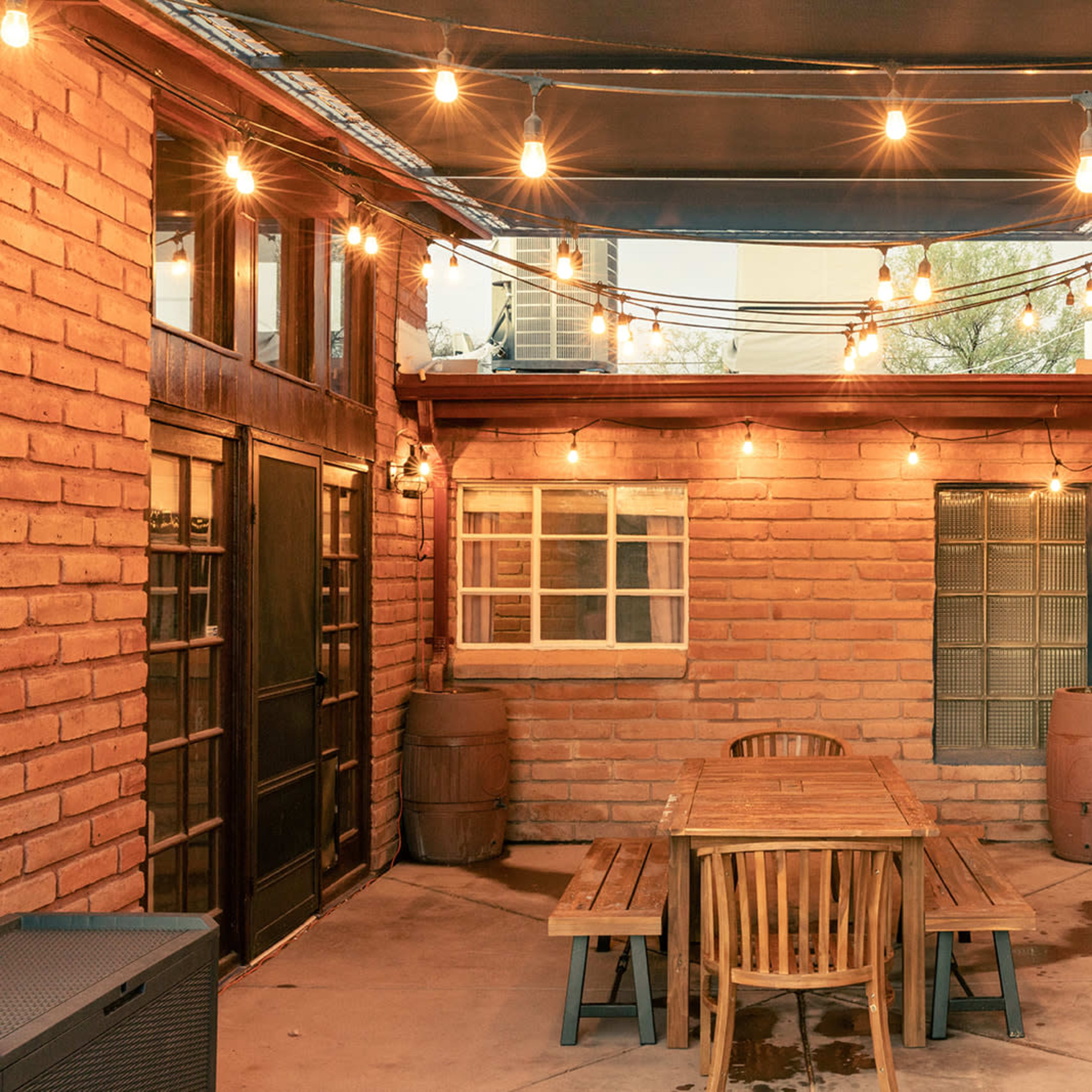 A courtyard with a wooden dining table surrounded by chairs, illuminated by string lights and brick walls.