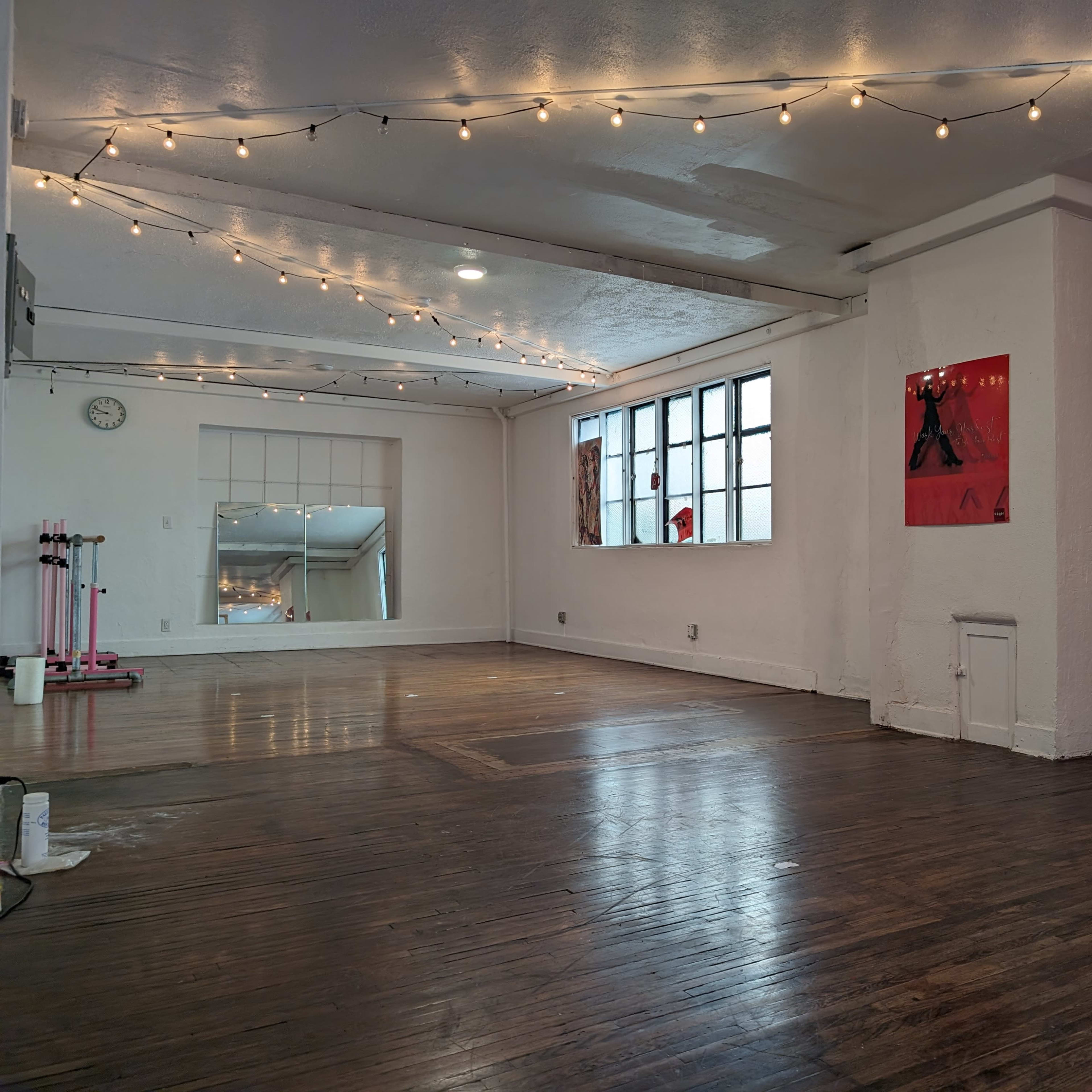 A spacious, well-lit room with wooden floors, large windows, and string lights overhead, alongside a brick-accented wall.